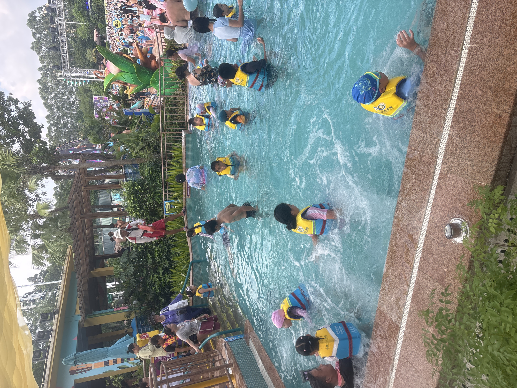 This is a daytime, eye-level shot of a crowded, outdoor swimming pool at a water park in Panyu District, China. The water is a clear blue, with visible ripples and splashes indicating activity. Numerous people, predominantly children wearing bright yellow life vests, are dispersed throughout the pool. Some are standing, while others are swimming or playing in the water. Adults are also present, some supervising, and others participating. In the background, a lifeguard in a red and white uniform stands by the edge of the pool. Beyond the pool, the water park environment is visible, with palm trees, lush greenery, water slides, and structures suggesting entertainment and amenities. The overall atmosphere is one of recreation and enjoyment.