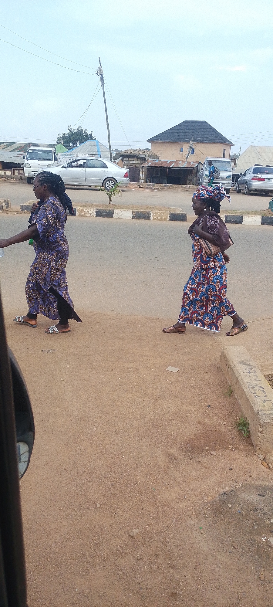 Two women are walking on a dusty road in Nigeria. The woman on the left wears a long, blue and brown patterned dress and black leggings, with her hair in braids. She carries a green bottle in her right hand. The woman on the right wears a colorful patterned dress and a headscarf, with a baby wrapped and carried on her back. They are walking on the right side of the road, in the same direction.
The setting appears to be a semi-urban area with buildings and parked vehicles in the background. A silver sedan is parked on the left side of the road, and a white van is visible behind it. Further down the road, there are more buildings with corrugated metal roofs, and a larger building with a dark, tiled roof. A utility pole with wires runs across the sky. The sky is overcast, suggesting it might be a cloudy day.
The ground is a mix of dirt and gravel, with a concrete curb running along the edge of the road. The overall impression is of a normal day in a Nigerian town, with people going about their daily activities. There are no clear indications of time of day other than the light level suggesting daytime. The emotion conveyed is neutral, simply two individuals walking. No visible text is discernable in the image.