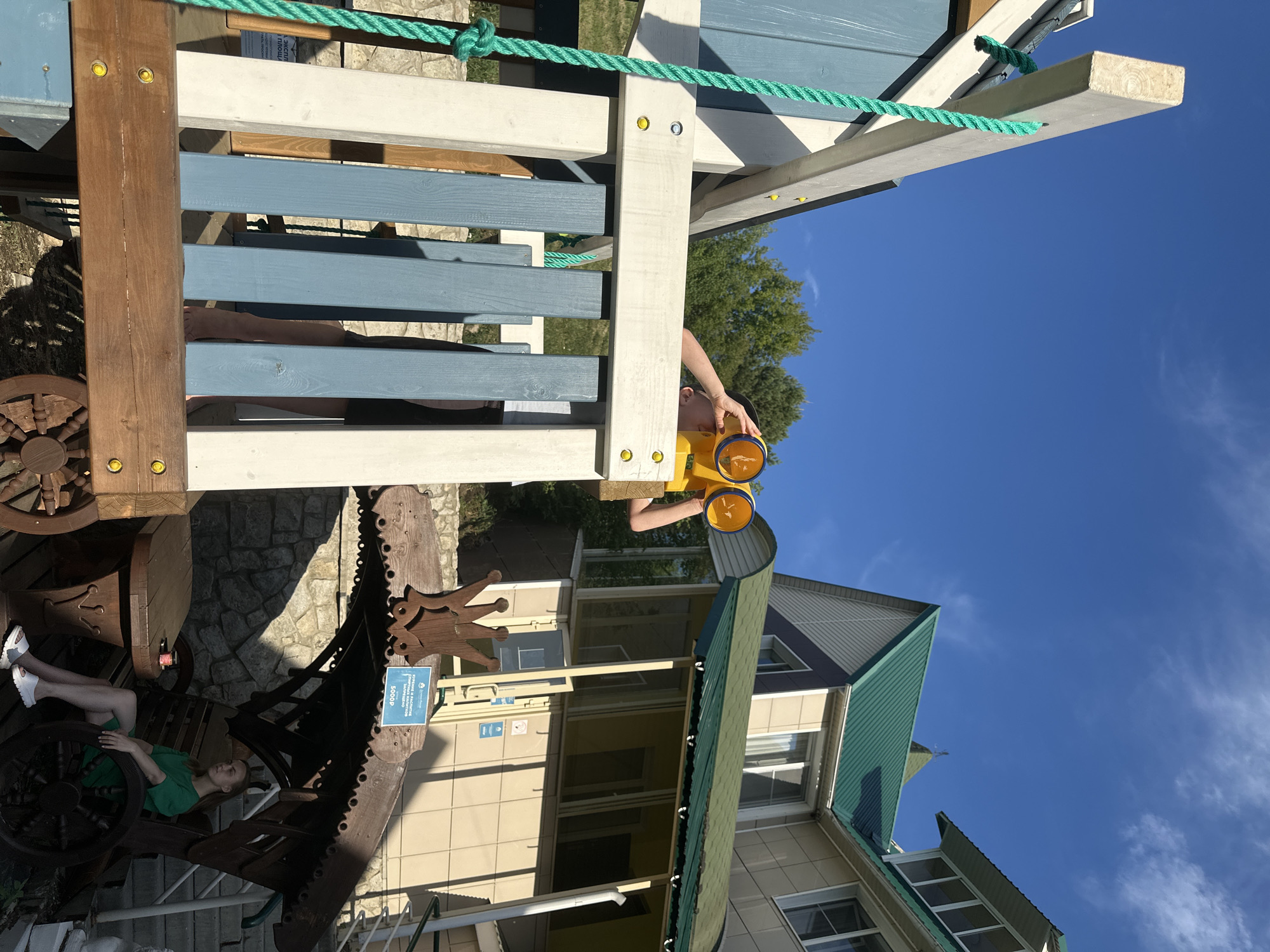 In the city of Danilovka (Altai), Russia, a young person, wearing a white t-shirt and dark shorts, stands on a wooden playground structure, looking through yellow binoculars. The playground features blue and white painted wooden planks and a green rope. In the background, a multi-story building with a green roof and beige siding is visible, along with trees and a clear blue sky. To the right of the playground, a young woman wearing a green dress sits on a wooden bench under a decorative wooden canopy. A stone wall and stairs are also present in the background.