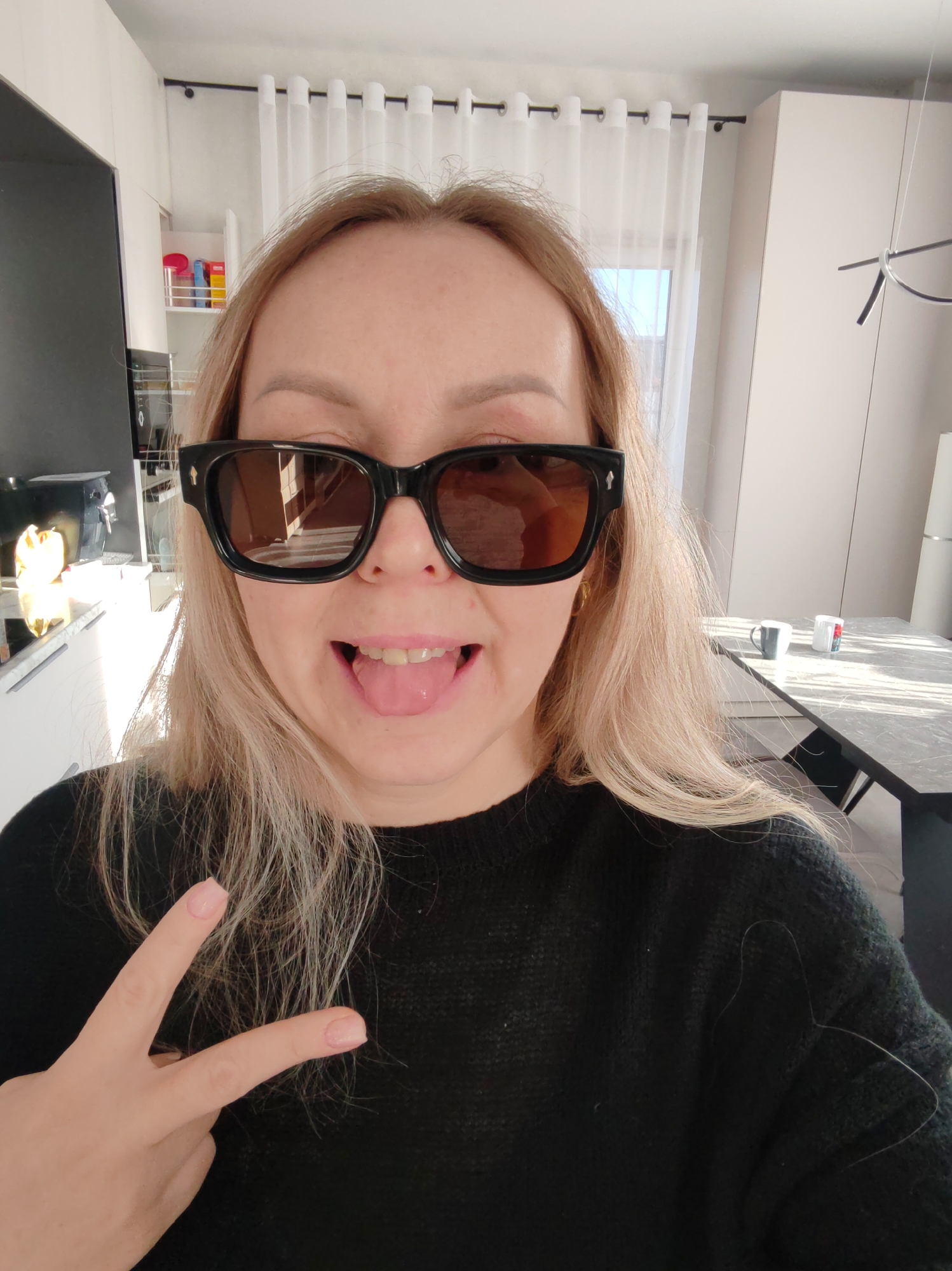 A fair-skinned woman with blonde hair is taking a selfie indoors. She is wearing large, dark-framed sunglasses and a black sweater. She is sticking her tongue out and giving a peace sign with her left hand. The background shows a kitchen area with white cabinets, a television, and a dining table with mugs on it. Curtains are visible in the window. There is no information available from the image to confirm the location.
