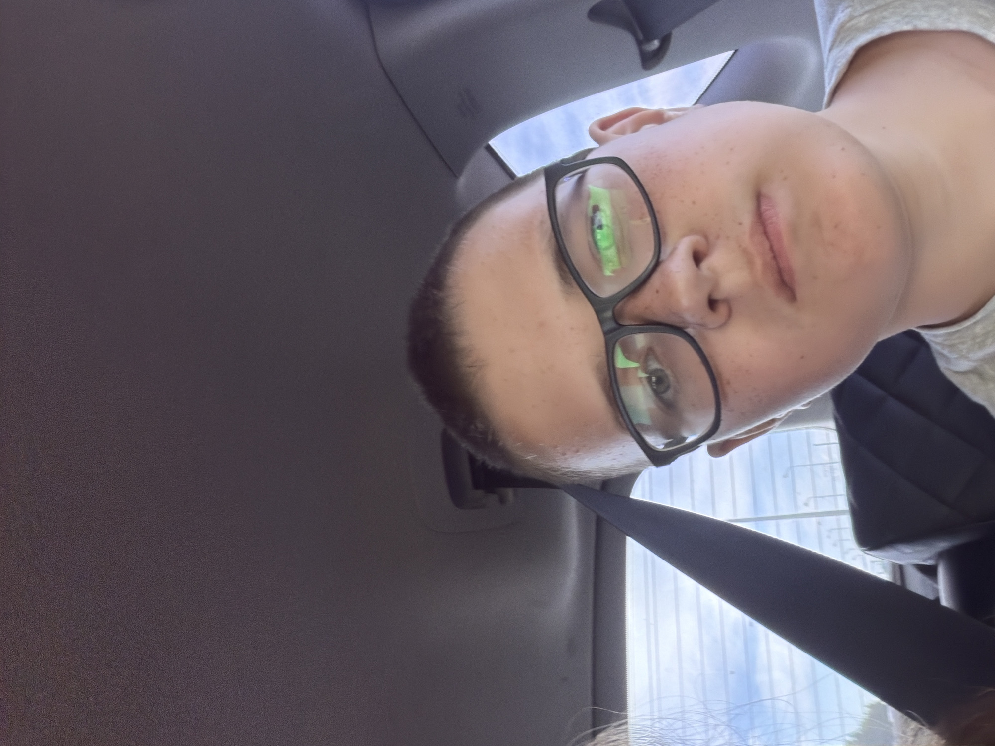 A young boy with short hair and glasses is taking a selfie from the passenger seat of a car. The boy is wearing a grey t-shirt and has freckles across his nose and cheeks. The car interior, including the headliner and a seatbelt, is visible in the background. Outside the car window, a cloudy sky and some distant trees can be seen. The boy's eyes are reflecting green light, suggesting the presence of a screen or bright lights. The car appears to be parked or moving slowly. There is no indication that this image was taken in the city of Новое Маврино, Russia.