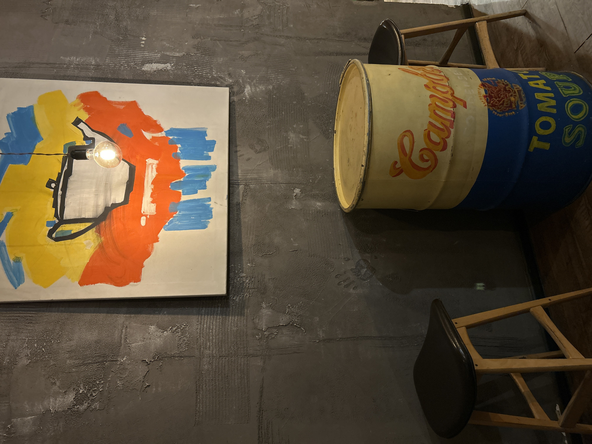 This media file captures an indoor scene with an industrial-chic aesthetic, likely a cafe or bar. The primary elements are set against a dark, textured gray wall, possibly concrete or a similarly finished surface, exhibiting visible trowel marks and some wear.
On the left side, a large, brightly colored abstract painting hangs on the wall. It features prominent areas of yellow, orange, and blue, with black outlines defining abstract shapes. An exposed incandescent light bulb, shining brightly, is suspended directly in front of the painting, creating a strong highlight.
To the right, a large metal barrel is mounted horizontally on the wall, designed to mimic a classic Campbell's Tomato Soup can. It displays the iconic yellow background with the red "Campbell's" script logo. A blue section of the barrel clearly shows the text "TOMATO SOUP" in yellow letters.
Below the barrel, two wooden bar stools with dark, possibly black, padded seats are partially visible. They are positioned as if against a counter or bar area, though these are not fully shown. The scene is illuminated by artificial light, and no people are present. While the location context is Saint Petersburg, Russia, no specific visual cues within the image confirm this.