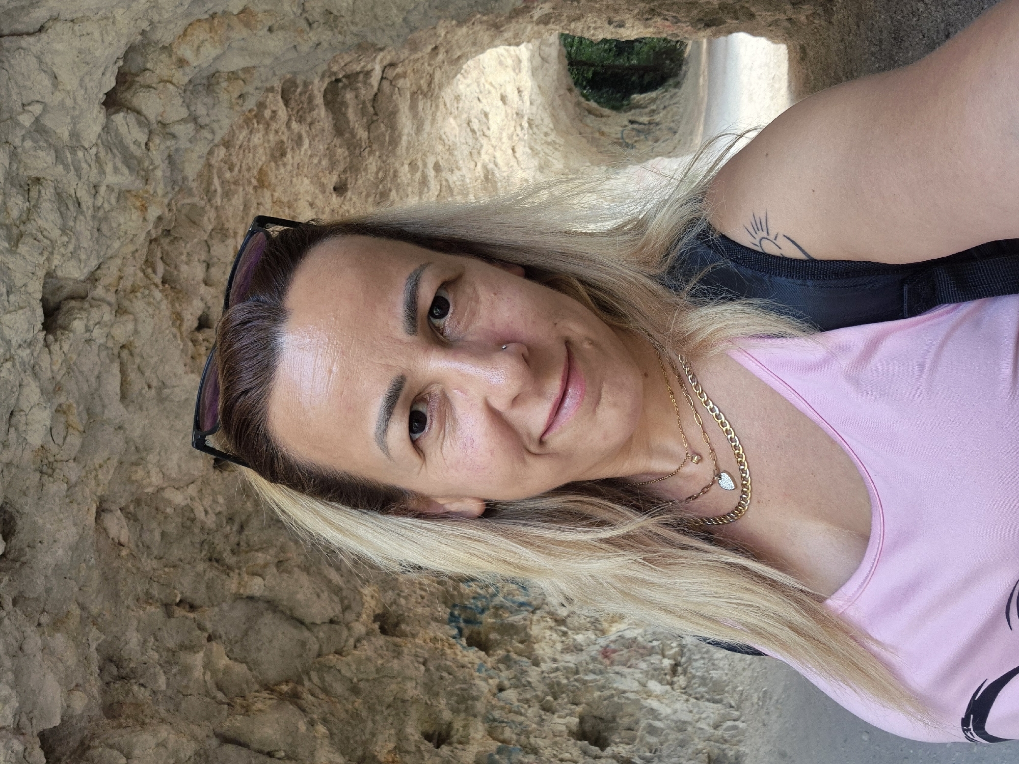 In a selfie taken in a cave-like natural tunnel with rough, light-brown rock walls, a woman with blonde hair pulled back and sunglasses on her head smiles at the camera. She wears a pink tank top, layered gold necklaces, and has a nose ring. A small sun tattoo is visible on her right shoulder. The tunnel extends into the background, with sunlight illuminating the path ahead and suggesting an outdoor environment.