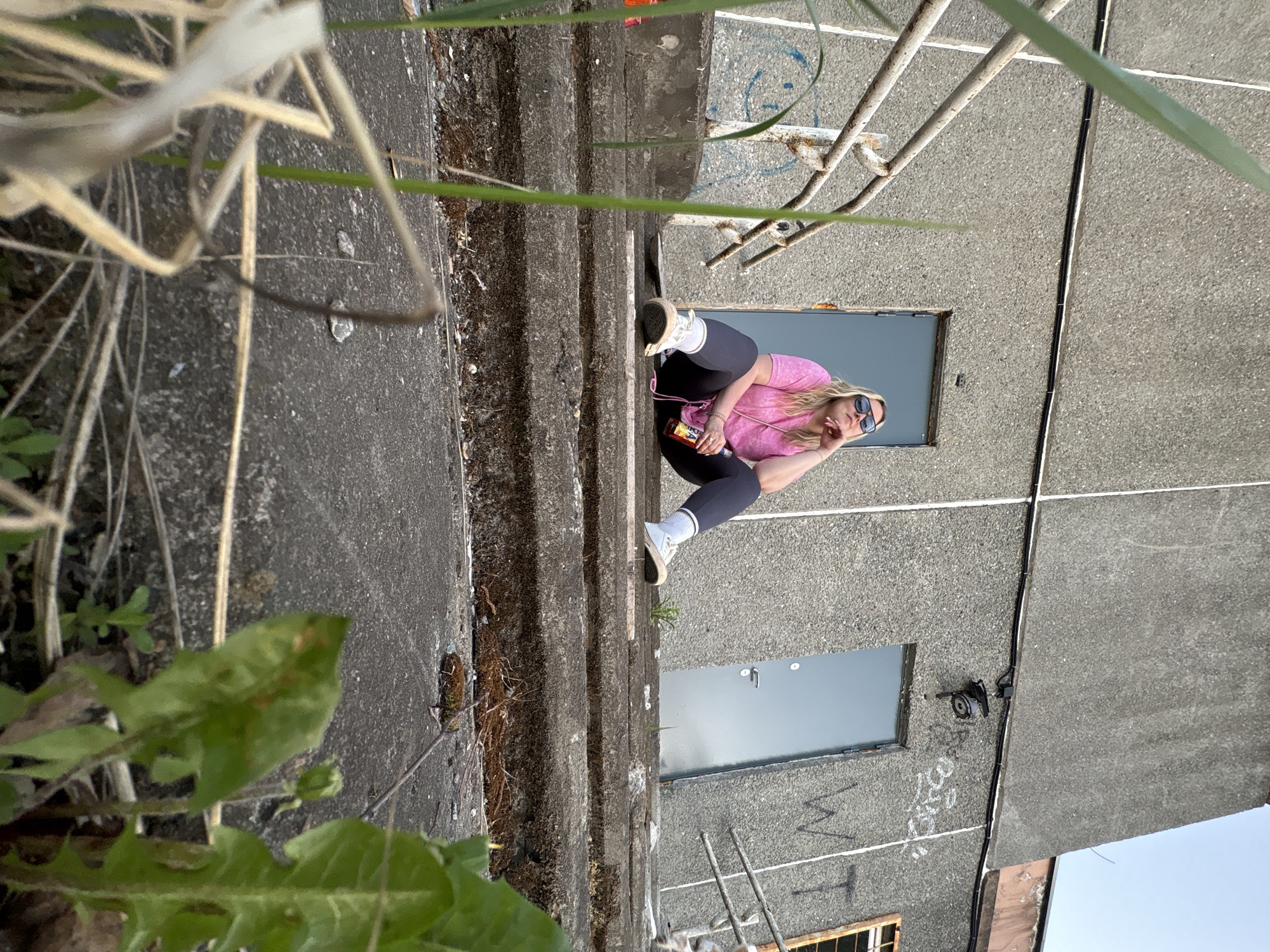 The image shows a woman in a pink shirt and leggings sitting on a concrete structure. She is wearing sunglasses and has a purse and a can next to her. The structure appears to be part of a building with two visible doors and graffiti on the exterior wall. There is vegetation growing near the concrete structure. The location is Марковский, Russia.