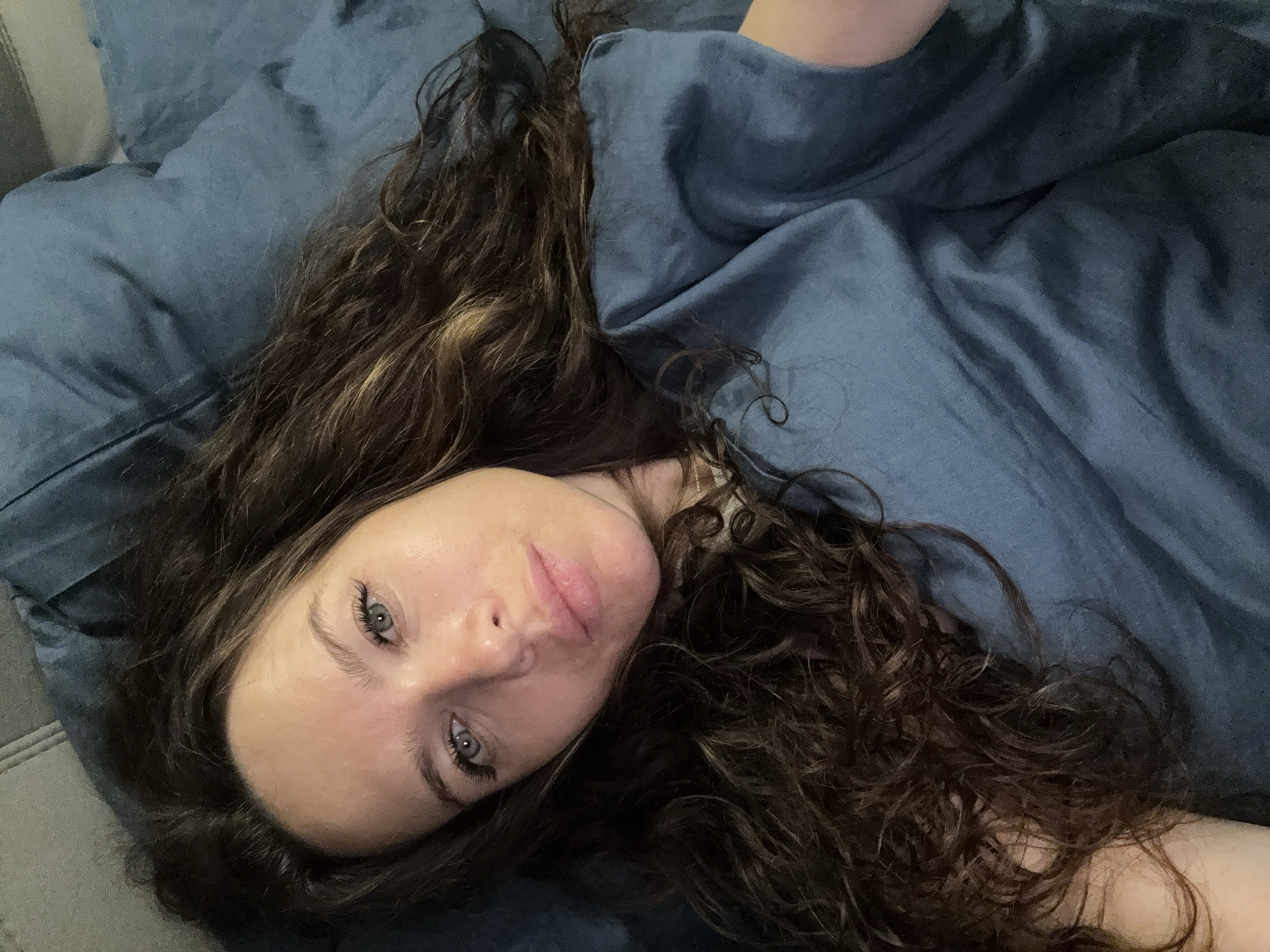A woman with long, wavy brown hair is lying on a blue surface. She has fair skin and light eyes. The background is a blue fabric, possibly a blanket or sheet. The location is not discernible from the image.