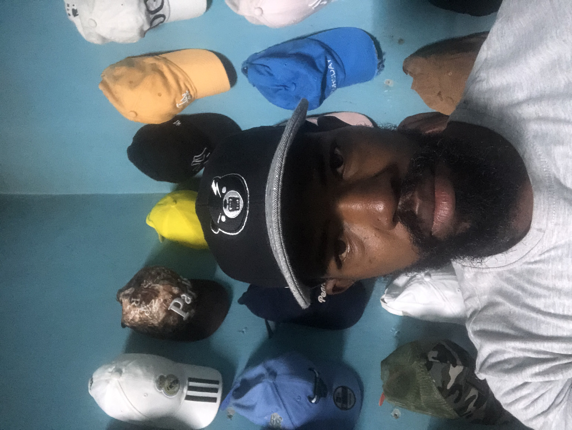 A man with a beard and a gray t-shirt is looking at the camera. He is wearing a black baseball cap with a white bear logo. The man is in front of a blue wall with many baseball caps hanging on it. The caps are of various colors and designs. Some of the caps are white, brown, yellow, black, and blue. There is also a camouflage cap. The scene is an indoor setting.