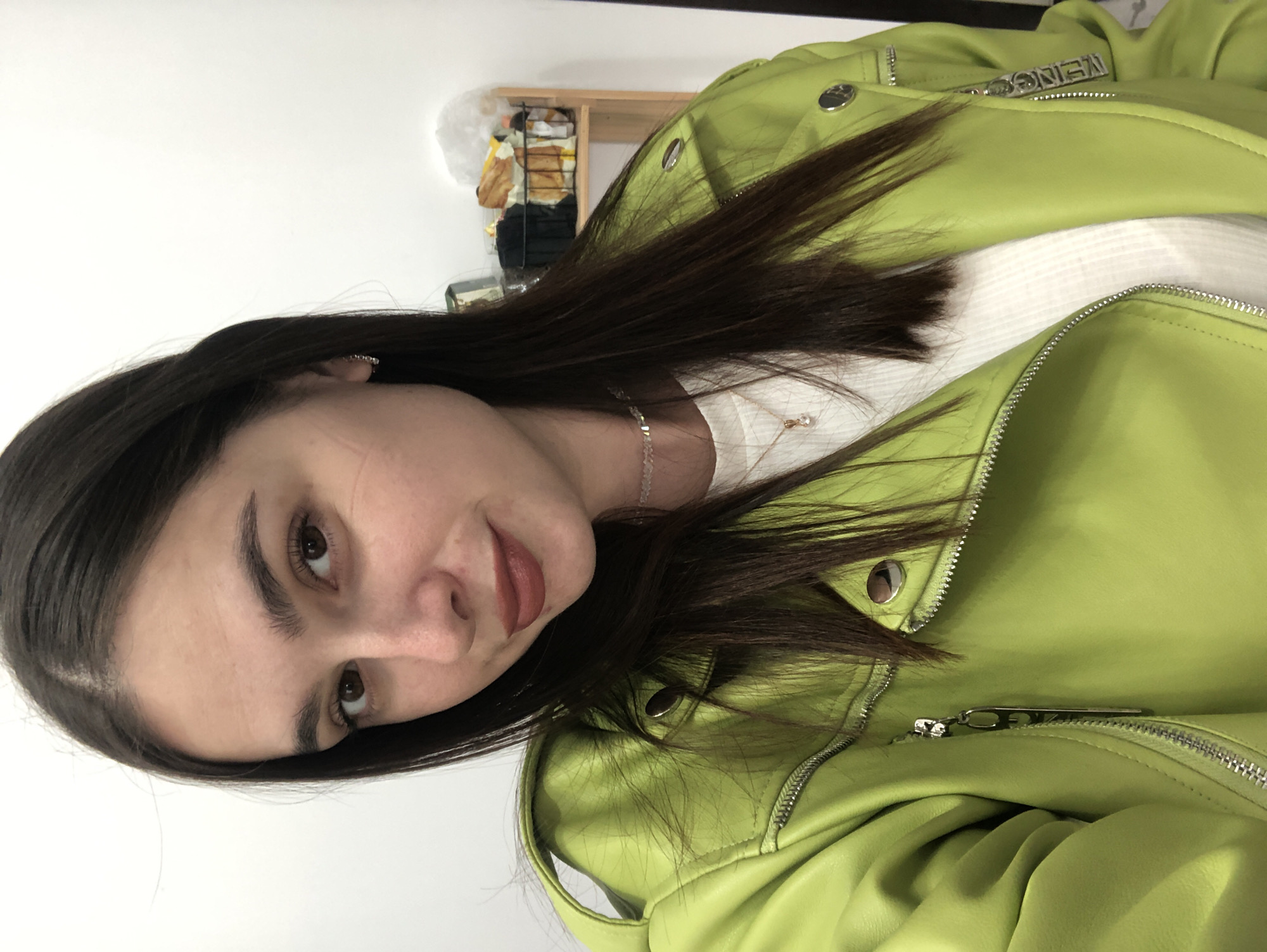 The media file features a close-up, overhead shot of a young woman with long, dark hair and brown eyes. She is wearing a bright lime green leather jacket over a white top and a delicate necklace with a small pendant. Her gaze is directed slightly to the left of the camera, and her lips are slightly parted, revealing a hint of a smile. The lighting suggests an indoor setting, likely during the daytime, with a plain white wall behind her. To the left of the frame, a shelf unit with various items is partially visible, including what appears to be some sort of packaging and decorative objects. There is some visible text on the zipper pull of her jacket which reads "NEINGO". The overall impression is that of a casual selfie or portrait.