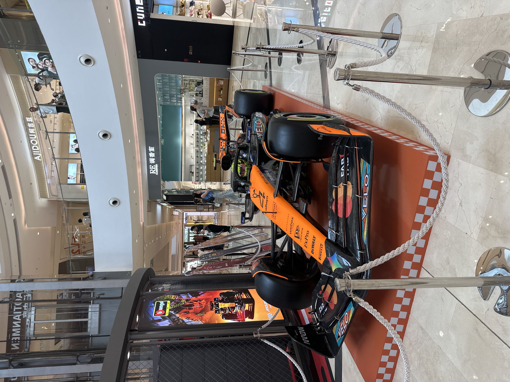 The image depicts an McLaren Formula 1 racing car, displayed within what appears to be a shopping mall or retail space in Yuzhong District, China, as indicated by Chinese signage present in the background. The car, featuring prominent orange and black colors, is positioned on an orange platform with a checkered border. A metal barricade, using ropes, separates the car from the public. Various logos and branding are visible on the car's body, including 'VELO', 'McLar', 'allwyn', 'Halton', 'webex', 'newera', 'FxPro', 'DEWALT', 'PIRELLI', 'RICHARD MILLE', 'DP WORLD' and 'FAR'. A screen displaying race car graphics sits near the base of the barricade. The surrounding environment includes retail stores, advertising displays, and pedestrian traffic. Overhead ceiling architecture with lighting fixtures is also visible.