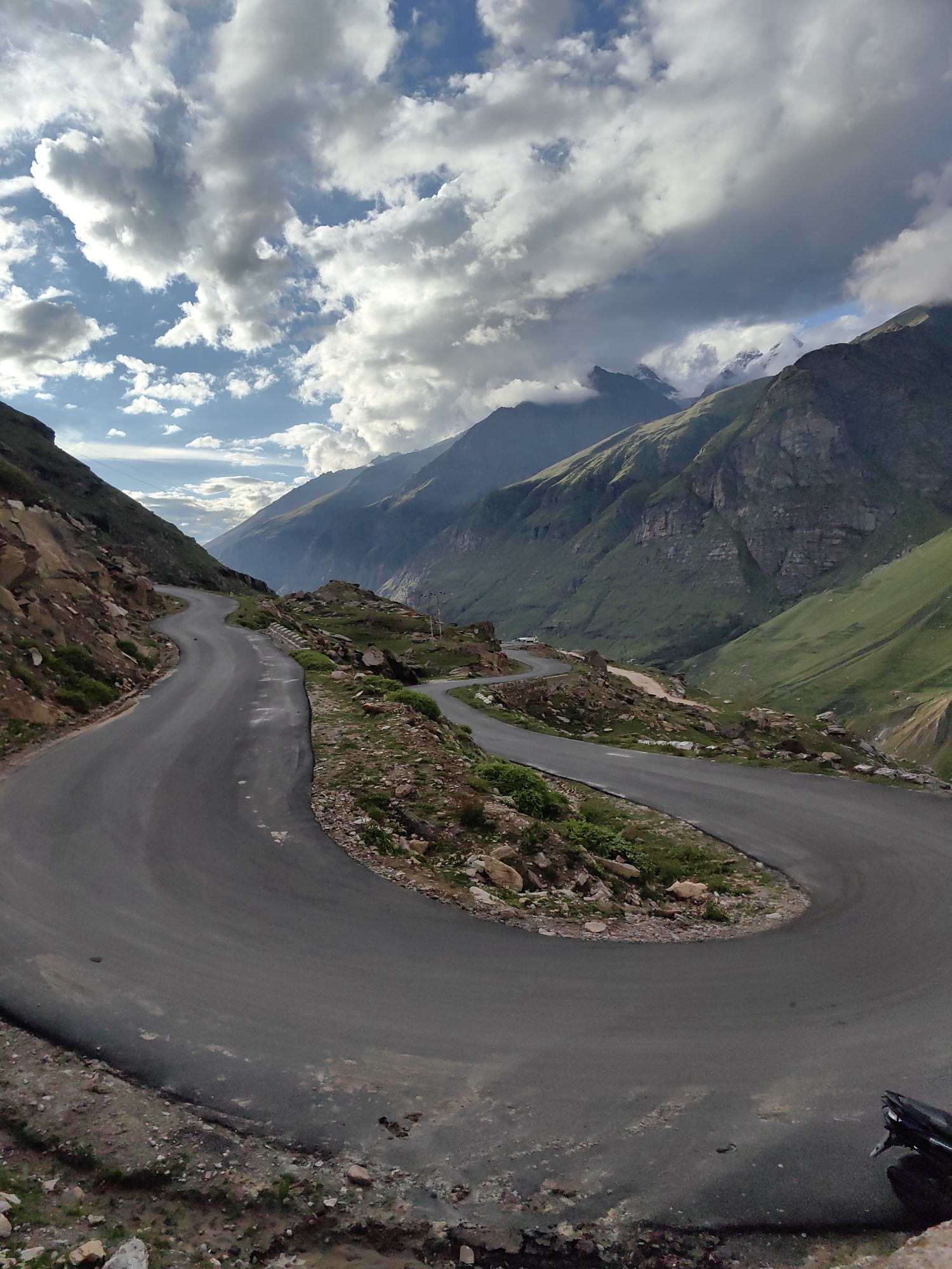 The image depicts a winding, paved mountain road in Gramphu, India, cutting through a dramatic landscape of steep, green slopes and rugged, rocky terrain. The road features several sharp hairpin turns, designed to navigate the challenging incline. The sky above is partially filled with dramatic, swirling clouds, with patches of blue sky visible, and sunlight intermittently illuminating the mountains. Distant, jagged peaks, some capped with snow or mist, rise in the background. The sides of the road are dotted with loose rocks and sparse vegetation, including small shrubs and grasses. A small section of a dark-colored motorcycle is visible in the lower right corner of the image. The overall impression is one of a remote and majestic mountain pass.