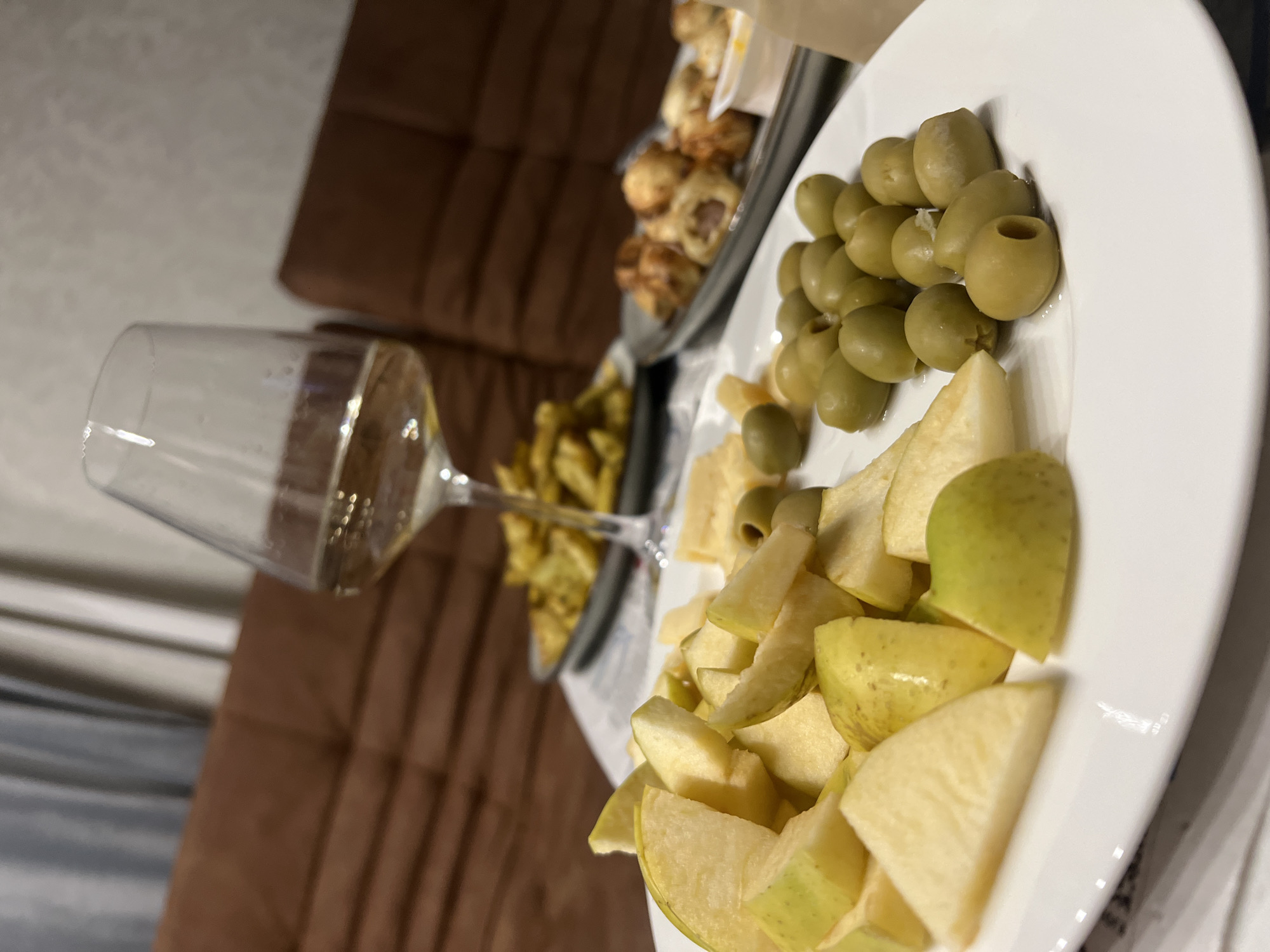 This image captures an indoor setting featuring an assortment of snacks and a drink, presented from a tilted, eye-level perspective. In the immediate foreground and to the right, a white, oval-shaped plate holds a substantial pile of green olives in the upper section and a generous heap of chopped green and yellow apple pieces in the lower section.
To the left of the plate, a clear stemmed glass, likely a wine glass or champagne flute, is positioned at an angle. It contains a light-colored, sparkling beverage, possibly white wine or champagne, with small bubbles visible within the liquid.
In the mid-ground, slightly out of focus, another serving dish or tray is visible, laden with several small, golden-brown baked items, which appear to be pastries or savory appetizers. The background features a blurred, brown, tufted or ribbed upholstered surface, consistent with a sofa or bench. Beyond this, a lighter, indistinct background element is present.
The lighting in the scene appears to be artificial, suggesting an indoor environment, possibly during the evening or night. The arrangement of food and drink indicates a casual gathering, an appetizer spread, or a relaxed snack time. No people are visible, and there is no discernible text within the frame. The overall atmosphere conveyed is one of casual enjoyment.