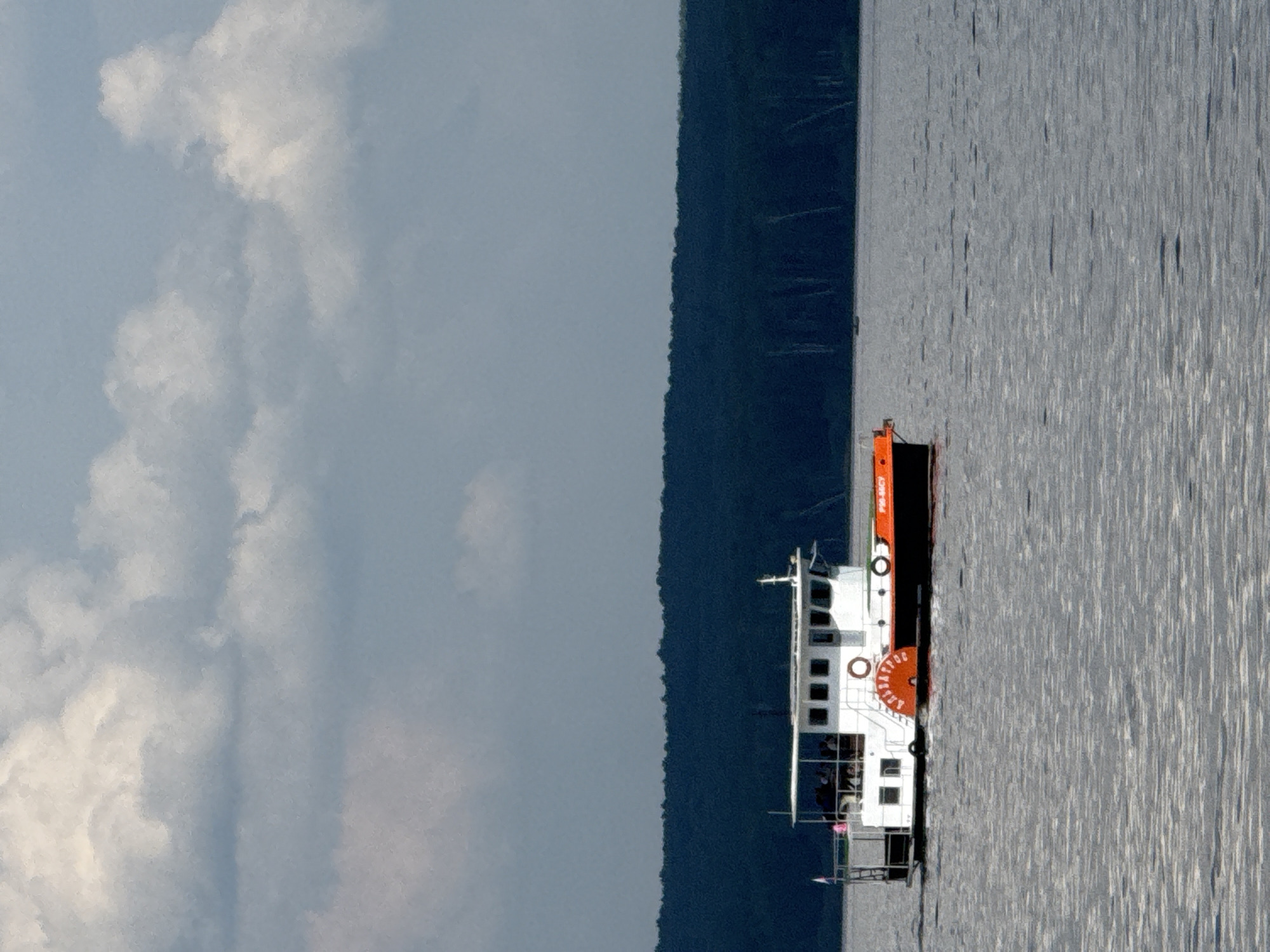 A steamboat cruises on the Kama River in Chaikovsky, Russia, beneath a cloudy sky. The boat is white with an orange hull, and a paddlewheel is visible on its side. Passengers are seated on the open deck. In the background, a dark, dense forest lines the riverbank. The water is calm, reflecting the overcast sky.