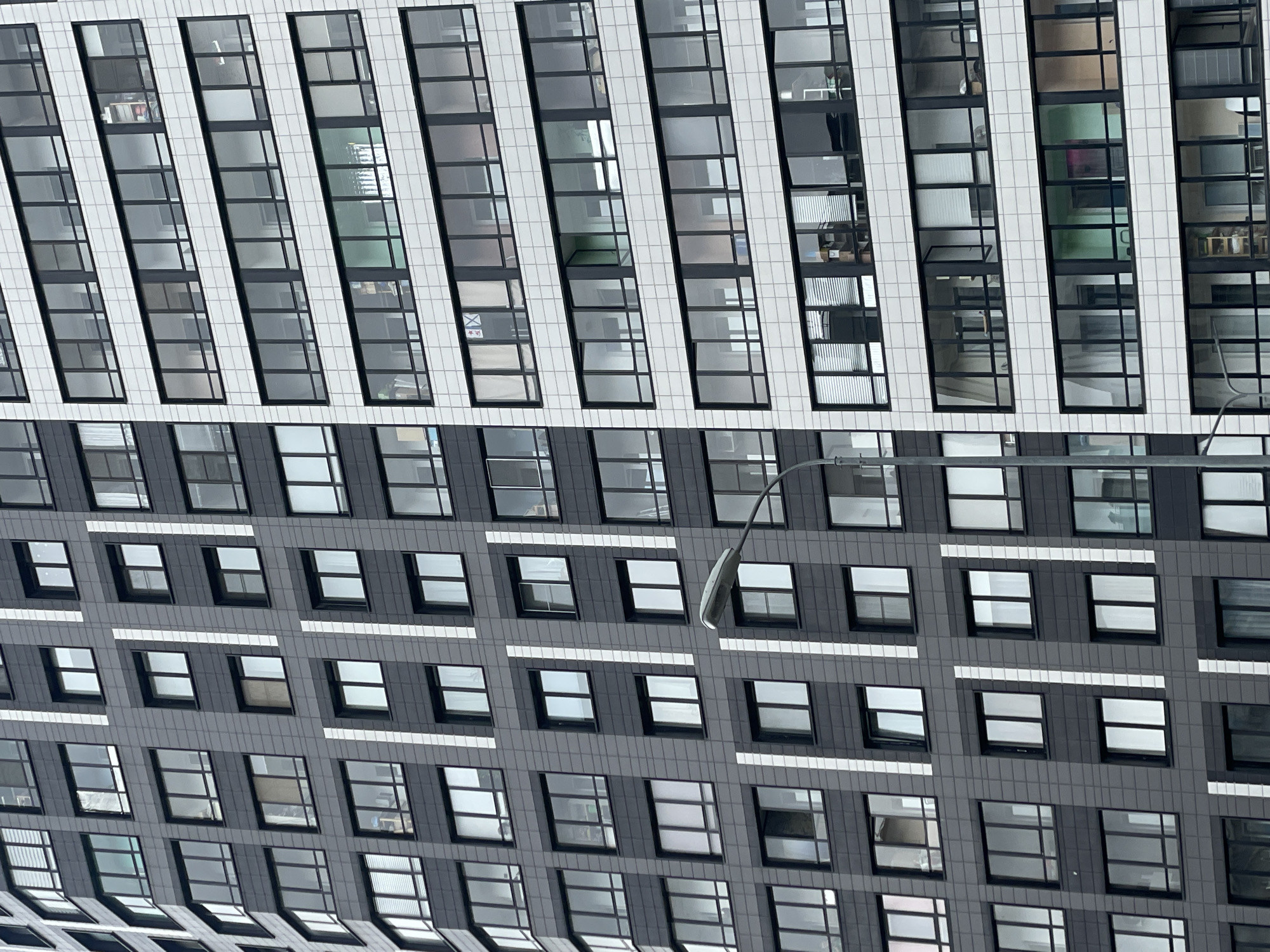 A modern high-rise building facade in Murino, Russia, is depicted in a vertically rotated shot, showcasing distinct architectural styles between its upper and lower sections.
The upper part of the building features a striking design of tall, narrow, and diagonally oriented window strips, framed in dark metal and alternating with textured white vertical panels. These windows often reflect an overcast sky, while some offer glimpses into interior spaces, revealing muted colors like light green, pinkish, or brown, and the indistinct shapes of office or residential furnishings. A small white sticker with a black 'X' mark and other indistinct symbols is visible on one of the lower windows within this diagonal section.
The lower two-thirds of the building presents a different aesthetic, characterized by a more conventional grid pattern of square and rectangular windows with dark frames, set within dark grey or black tiled panels. Horizontal white strips occasionally traverse this darker facade. Reflections in these lower windows vary, showing parts of the sky, interior lights, or other architectural elements. A prominent, curved street lamp arm with a modern light fixture extends into the frame from the right, partially obscuring the middle section of the lower facade.
The overall lighting suggests an overcast or cloudy daytime, providing diffuse illumination without harsh shadows, highlighting the building's textural and tonal contrasts. No people or direct activities are visible.