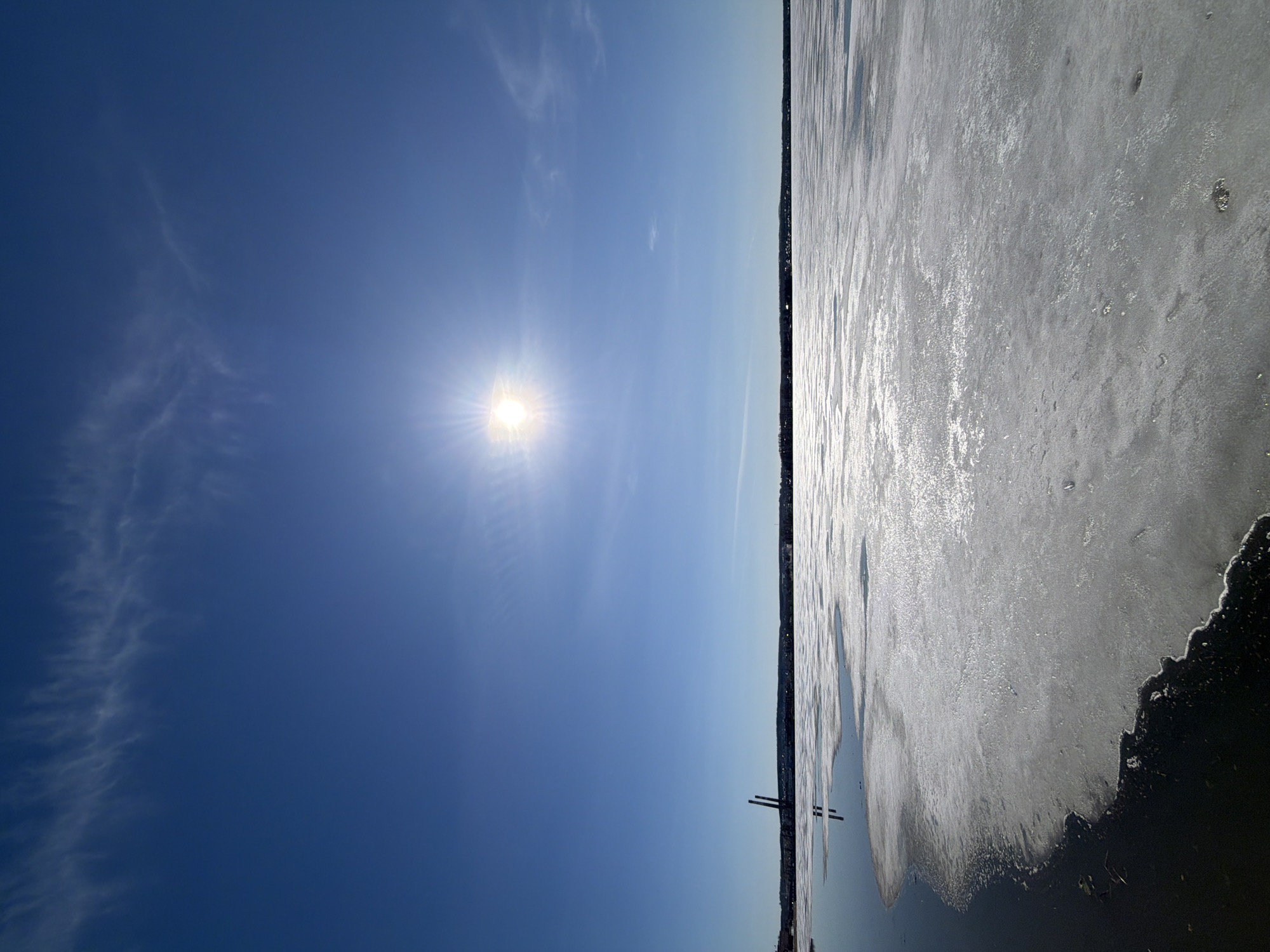 A landscape photograph captures a vast, partially frozen body of water under a bright blue sky. The sky, occupying the upper two-thirds of the frame, is a deep, clear blue with wispy white cirrus clouds scattered across it, particularly on the left. The sun shines intensely from the upper left quadrant, creating a prominent starburst effect and casting a strong glare.
The lower third of the image features a wide expanse of water, largely covered in ice. The ice surface appears somewhat slushy or broken, highly reflective, and glimmers brightly under the sunlight. Towards the right side of the frame, a darker area of open water or less reflective ice is visible. A thin, dark line on the horizon indicates a distant shoreline or landmass. Against this backdrop, two dark, slender, vertical structures, possibly poles or remnants of a pier, stand in the water near the center-right. The scene suggests a clear, sunny day, likely during late winter or early spring, given the state of the ice. No text is visible in the image. The setting is Votkinsk, Russia.