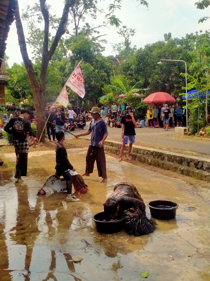 The image depicts an outdoor scene in Gebyog, Indonesia, likely a public event or performance, occurring on a muddy and wet ground surface with standing puddles. Lush green trees and foliage form the background, with a paved road and stone retaining wall visible on the right. A crowd of onlookers is gathered in the midground and background.
In the foreground, five males are present. From left to right:
1. A male, barefoot, wears a black long-sleeved shirt with a white graphic and dark checkered sarong/pants. He stands in a puddle, holding a long, thin stick or whip-like object.
2. A younger male, wearing a black long-sleeved shirt and dark pants with light-colored shoes, sits astride a white, stylized horse-shaped prop (likely for *Kuda Lumping*) which rests on the muddy ground.
3. A male, barefoot, wears a blue short-sleeved shirt, dark pants, and a light-colored hat. He stands in a puddle, extending a green plant stem or thin branch towards the male on the horse prop.
4. A male, barefoot, wears a black t-shirt with "BALINESE" in white text and blue shorts. He stands with his right foot lifted, observing the scene.
5. In the lower right, a male wearing a dark long-sleeved shirt with white patterns is hunched over, with his face submerged in a black plastic basin filled with dark liquid and a dark, feathery object. An identical empty black basin rests beside the first.
Two white flags with red text, one prominently displaying "REWANG," are visible in the midground, attached to poles. A red umbrella and a blue tent are situated further back on the right side of the road, near the spectators. A street light pole is also visible on the right.