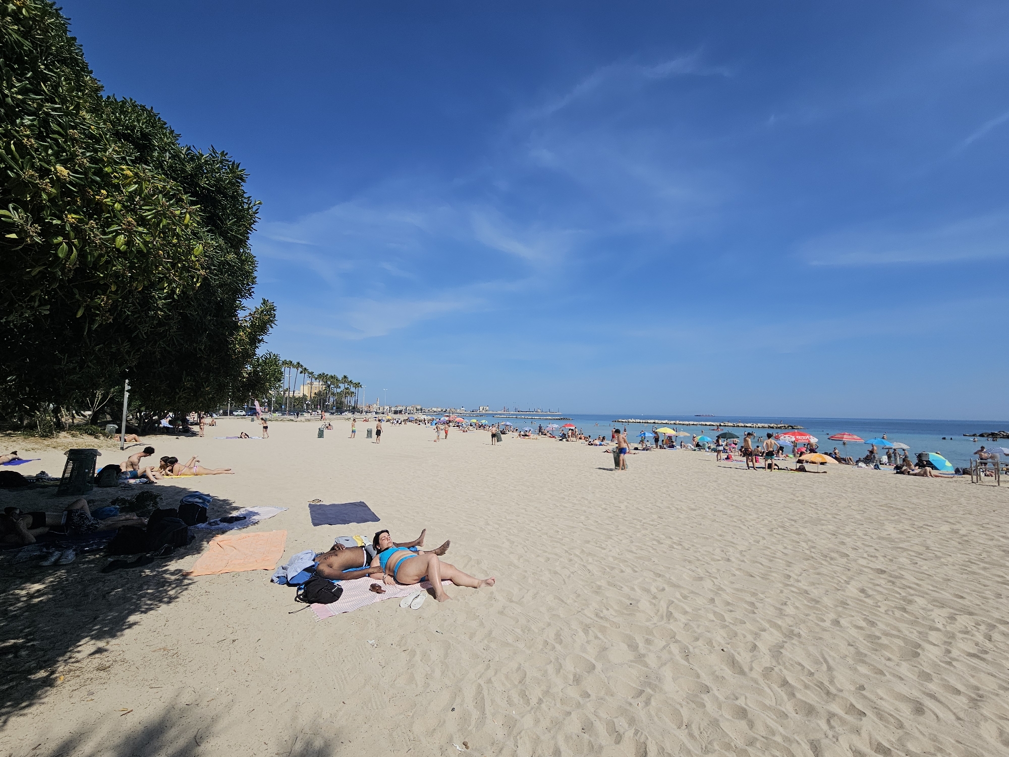 This image shows a beach scene in Bari, Italy, on a sunny day. The beach is sandy and crowded with people. Some are sunbathing, swimming, walking, and relaxing under umbrellas. A few individuals are lying on towels in the foreground. The background includes the sea and the skyline of Bari. A tree is on the left-hand side of the frame, with its shade cast on the sand. The sky is blue with scattered clouds.
