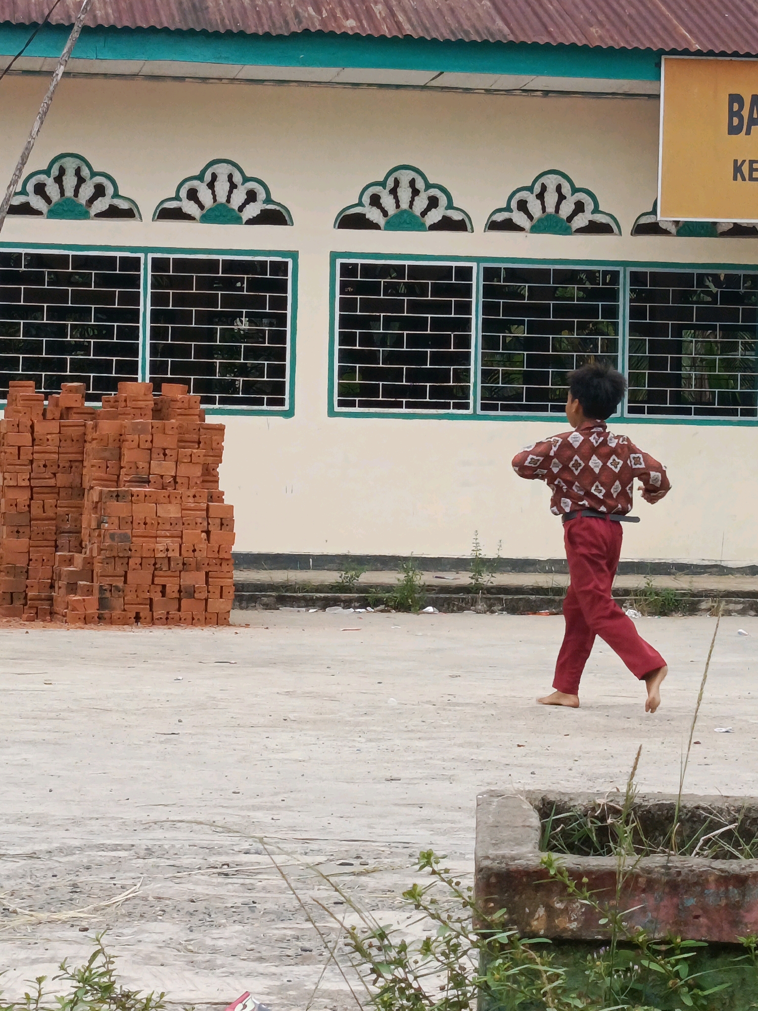 In the city of Wonosari, Indonesia, a young boy with dark, curly hair, wearing a red patterned shirt and red pants, walks barefoot across a dusty, concrete ground. He is captured mid-stride, his body turned slightly away from the camera, with his right arm bent and his left arm extended behind him, suggesting movement. To his left, a tall stack of red bricks is piled high, with some scattered on the ground around it. Behind the bricks and the boy, a light-colored building stands with a dark green trim and a red, corrugated metal roof. The building features decorative white and green arches above windows that are covered with metal grates. A yellow sign with black lettering is visible on the right side of the building. The environment appears to be an outdoor space, possibly a construction site or schoolyard, with sparse vegetation and some litter on the ground.