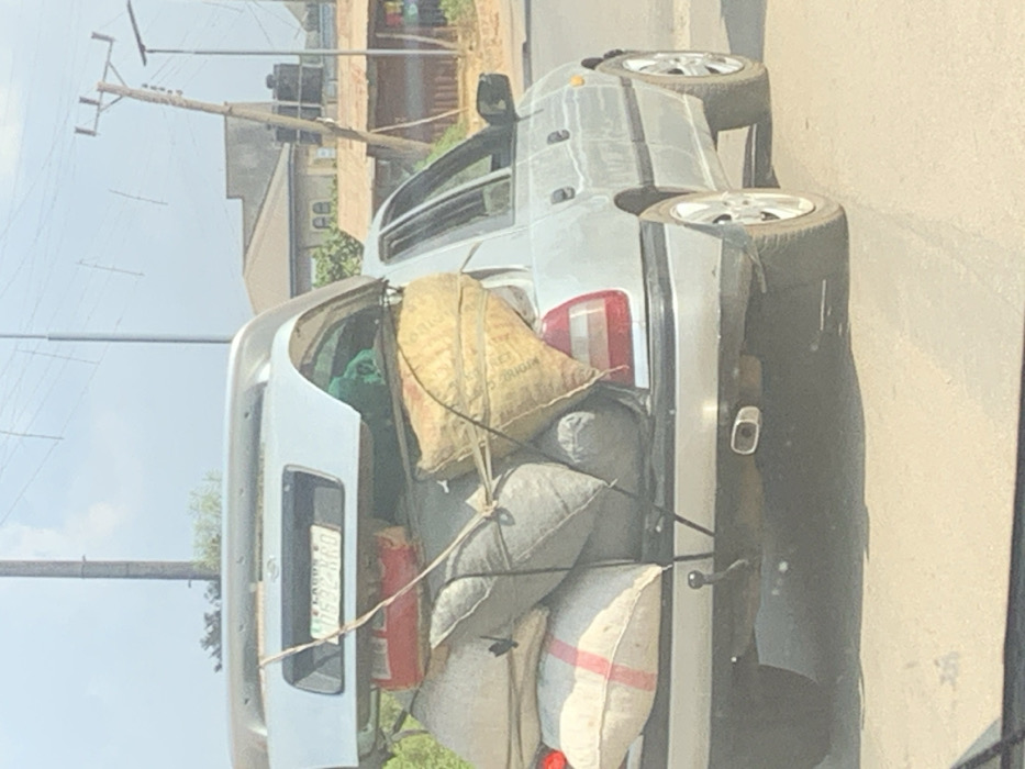 A silver sedan, likely a Nissan model, is depicted from a rear-quarter perspective on a bright day in Egbedi, Nigeria. The vehicle is stationary or moving slowly on an asphalt surface, heavily overloaded in the trunk area. The car's original trunk lid is detached and positioned horizontally on top of the stacked cargo, displaying a Nigerian license plate "LAGOS 2 D532 KRO." The open trunk is filled with numerous large, irregularly shaped sacks and bags; colors include light grey, beige (one with visible text), and white with a red stripe. A red rectangular container and green fabric are also visible amidst the load. Manila-colored ropes secure the cargo and the detached trunk lid to the vehicle. The car's rear features a single exhaust pipe and a tow hitch. In the background, under a clear sky, are utility poles with overhead power lines, residential-style buildings with light-colored roofs, and a smaller structure with a corrugated metal roof and visible black water tanks. No individuals are discernible within the frame.