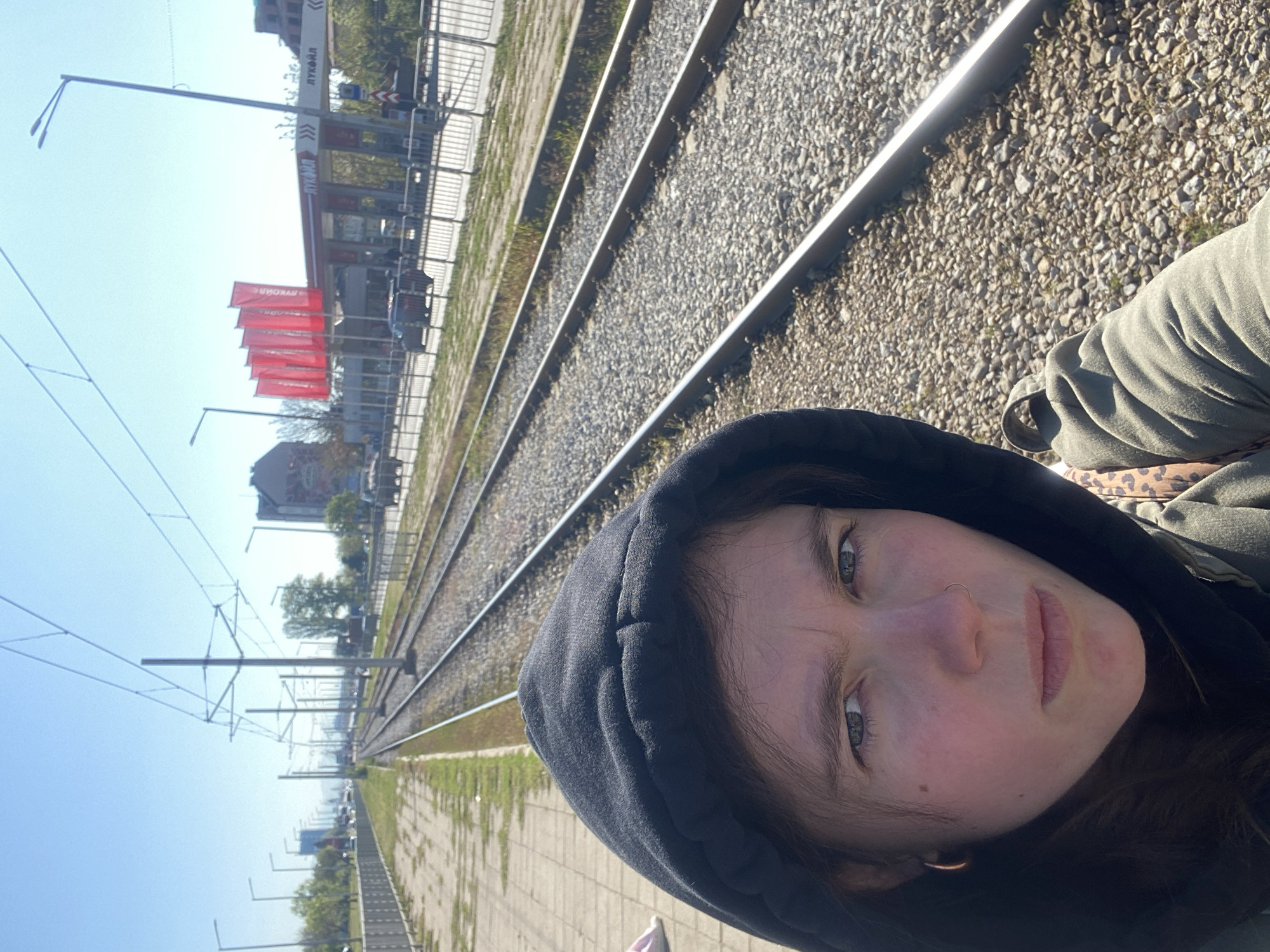 A selfie shows a woman wearing a black hooded sweatshirt near train tracks. The location is in Sofia, Bulgaria, identifiable by the ЛУКОИЛ gas station visible in the background. The scene includes train tracks covered with gravel, concrete tiles on the ground, and overhead electrical wires for the trains. The woman is the primary subject, with the train tracks and gas station forming the background. The sky is clear.