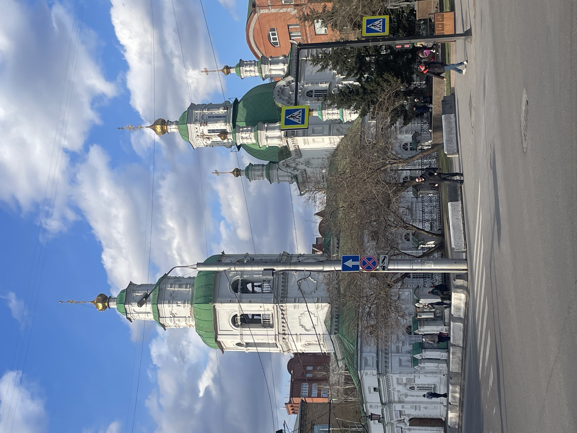 The image shows a street scene in Krasnoyarsk, Russia, featuring the Annunciation Cathedral prominently on the left. The cathedral has white walls, green roofs, and gold domes. Bare trees stand in front of the cathedral along the street. A few people are walking or standing along the sidewalk on the right side of the image, which runs alongside the road. There are crosswalks marked on the road. Various traffic signs are visible, including pedestrian crossing signs and no parking signs. Buildings are visible on both sides of the road. Overhead power lines crisscross the sky, which is partially covered with clouds.