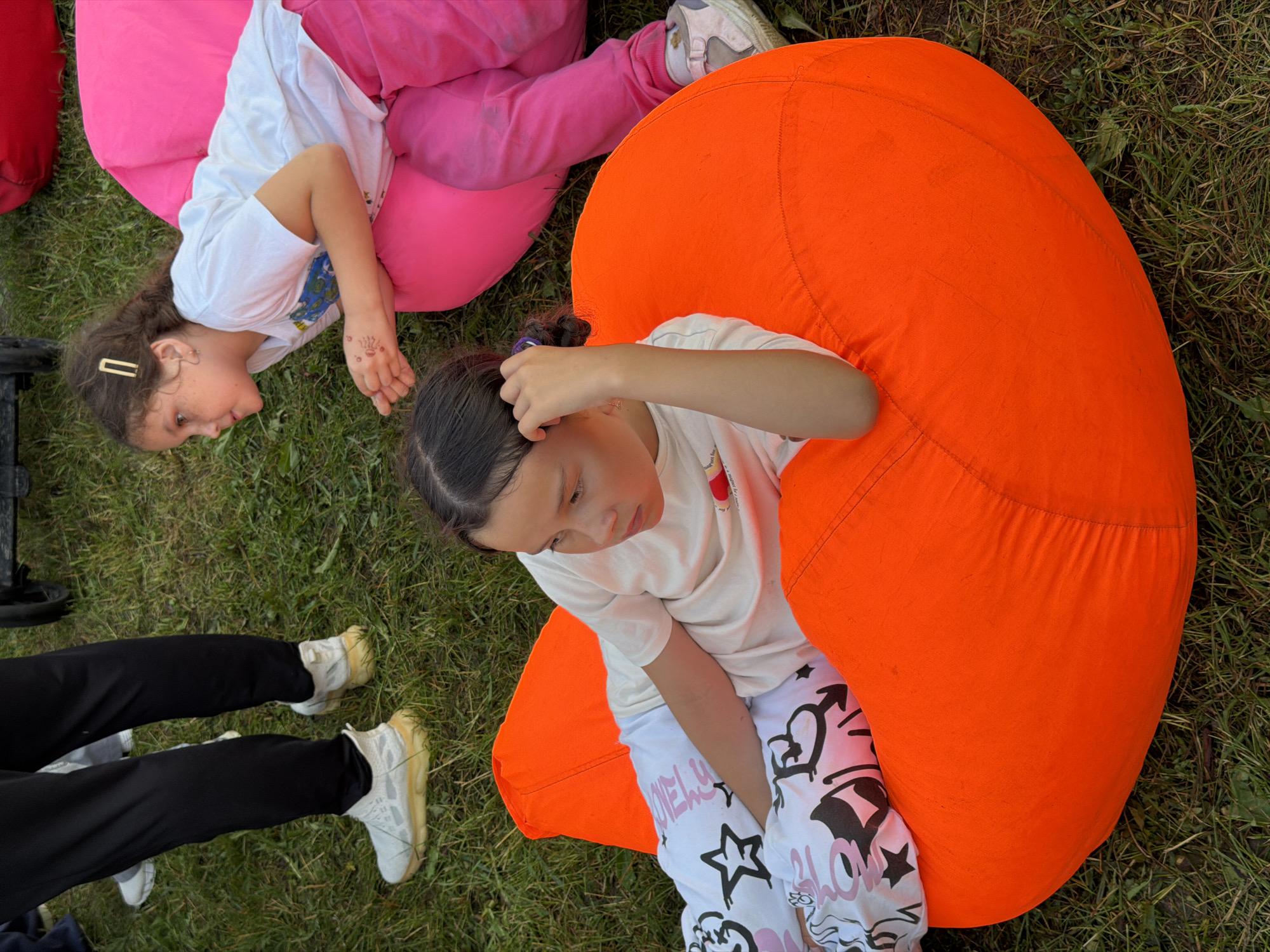 In an overhead shot, two young girls are seen lounging on beanbag chairs in an outdoor grassy area. One girl, on the left, is lying on a large pink beanbag chair, dressed in a white t-shirt and pink pants. She has a gold hair clip in her brown hair and what appears to be henna markings on her right hand. Her gaze is directed downwards and to her left.
The second girl is positioned on a bright orange beanbag chair, situated to the right and slightly above the first girl in the frame. She is wearing a white t-shirt and white pants adorned with black graffiti-style drawings, including hearts and stars, and the word "BLOW" is visible. She is touching her hair with her right hand and looking down towards her pants.
The scene suggests a relaxed, informal gathering. The presence of beanbag chairs and the natural setting imply an outdoor event or leisure time. The lighting indicates it is daytime. The overall mood appears to be peaceful and casual, with the children seemingly comfortable and engaged in their individual activities.