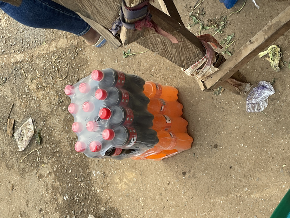 A pack of bottled soda sits on a rough, dirty ground. The pack consists of Coca-Cola bottles with red caps and orange-colored BRC soda. A wooden structure, possibly a makeshift stand, is partially visible near the soda. There are also pieces of trash and debris scattered around. A person wearing jeans and sandals is seen in the top left corner, likely standing near the drinks.