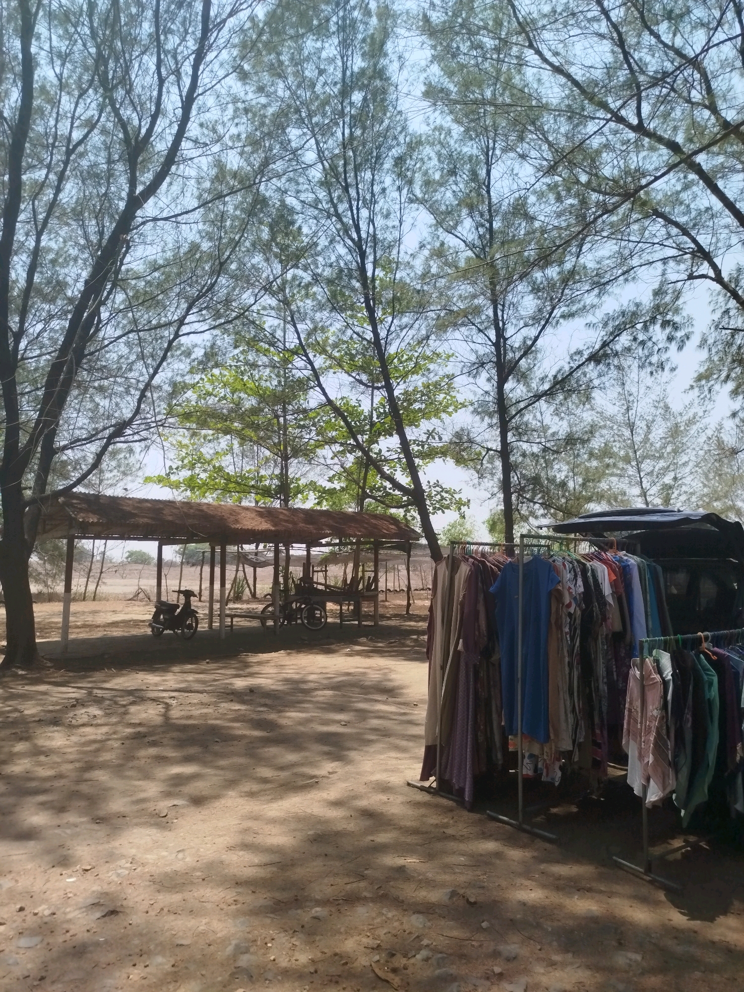 This image depicts an outdoor stall selling clothing, likely in Buluspesantren, Indonesia. The stall features racks of colorful garments, including dresses, shirts, and scarves, displayed in front of a dark-colored vehicle. In the background, a simple open-air shelter with a thatched roof covers a motorcycle and a wooden cart. The ground is dusty and uneven, with dappled sunlight casting shadows from the surrounding trees. The overall environment suggests a rural or semi-rural setting.