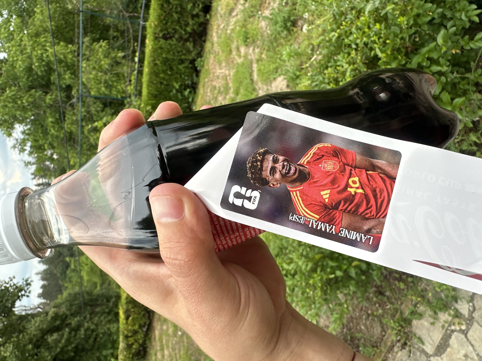 A hand holds a dark glass bottle of Coca-Cola with a soccer player's trading card attached to it. The card features a young man in a red and yellow soccer jersey, identified as "LAMINE YAMAL (ESP)". The background shows lush green foliage and trees, suggesting an outdoor setting. The image was taken in Prisovo, Bulgaria.