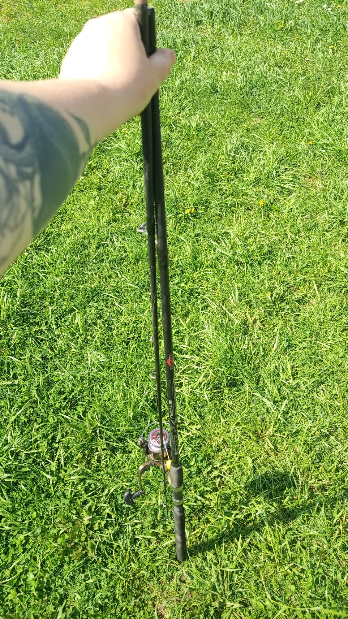 A person is holding a fishing rod in their right hand, standing on a patch of green grass. The rod is black with a silver reel. The person has a tattoo on their arm. The location is a grassy area, but there is no additional evidence to determine if it is in Уральское, Russia.