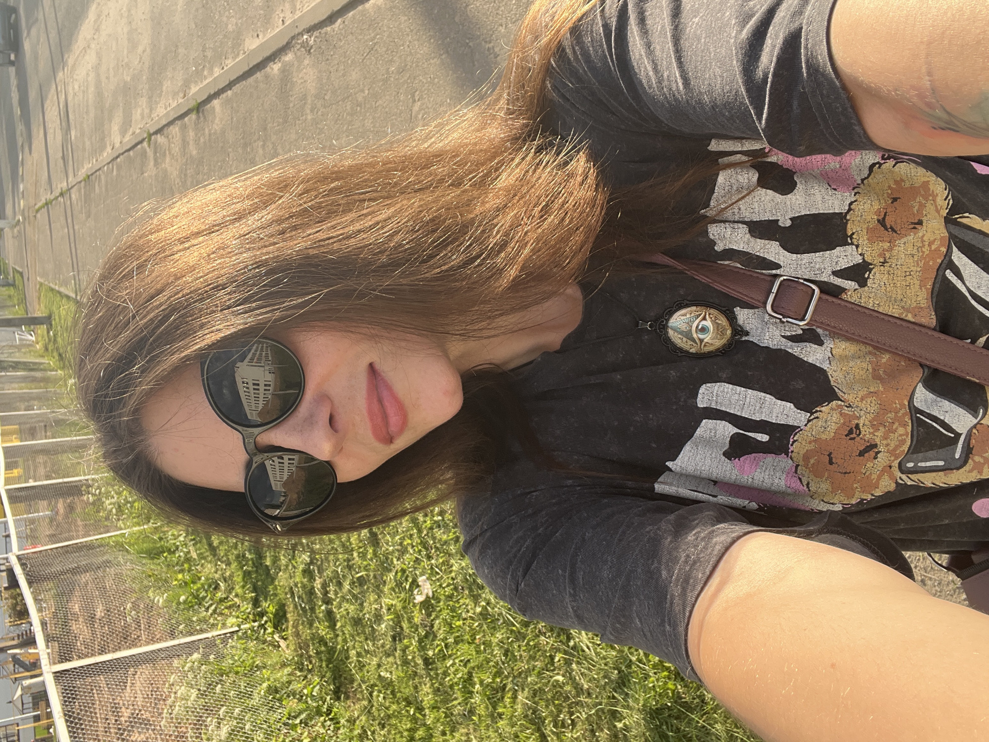 A woman with long brown hair, wearing sunglasses and a dark t-shirt with a graphic print and a pendant, takes a selfie outdoors. She is standing on a paved path next to a grassy area with a chain-link fence in the background. The sun is casting shadows, suggesting it is either morning or late afternoon. The reflection in her sunglasses shows a building. The image appears to have been taken in a residential or semi-rural area, possibly in the city of Krasnoe-na-Volge, Russia, given the context of the fenced-off area and the style of the fence.