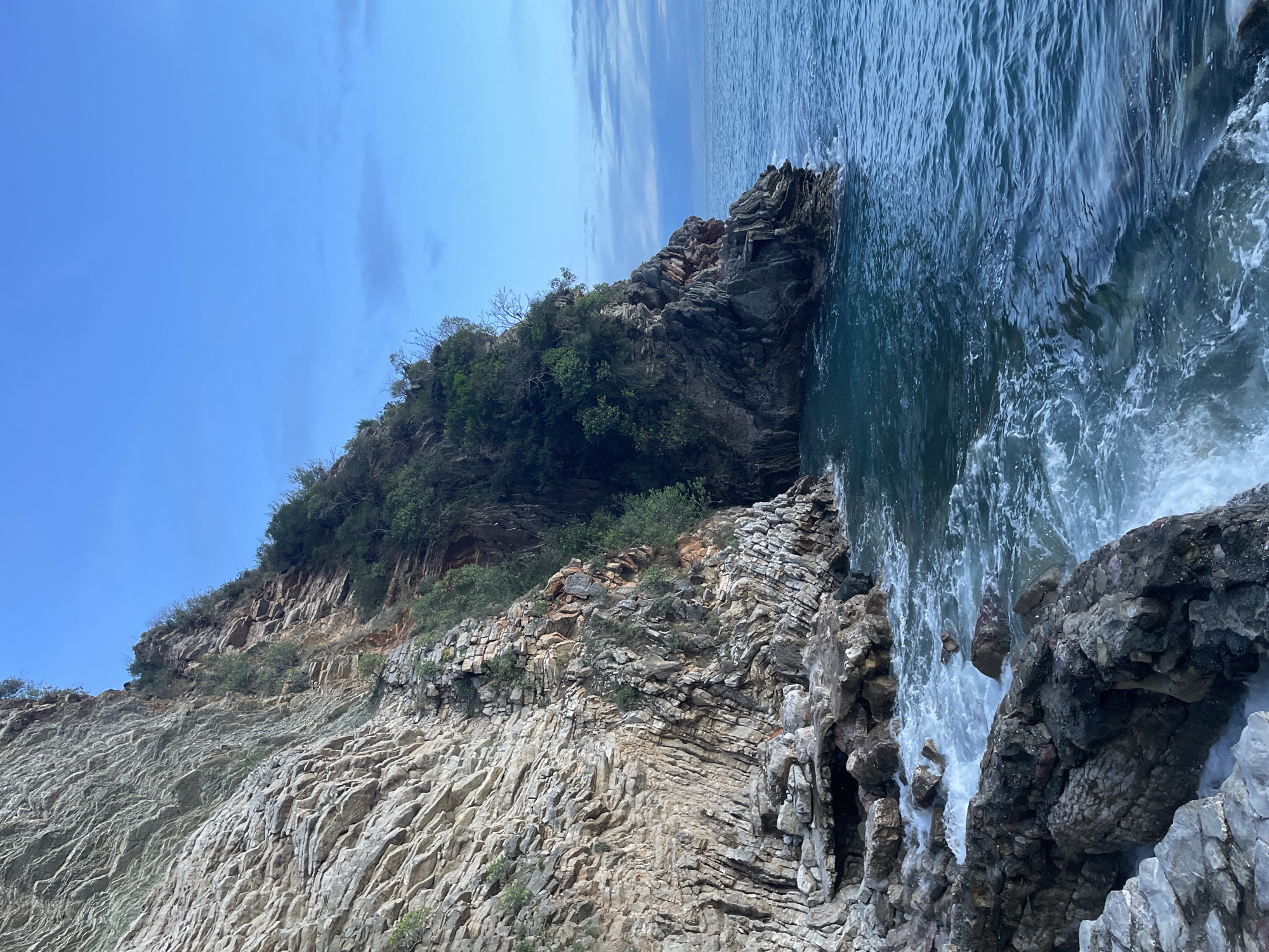 The image depicts a natural coastal scene in Rafailovići, Montenegro, focusing on a rugged cliff face meeting the sea. The left half of the frame is dominated by a towering, stratified cliff composed of light grey and tan sedimentary rock layers, exhibiting significant vertical and horizontal fracturing. Sparse green vegetation, consisting of small bushes and trees, grows from crevices and ledges along the cliff, becoming denser towards the top and on the right side of the rock formation.
The right side of the image features the deep blue-green waters of the Adriatic Sea. Waves are actively breaking against the base of the cliff and the dark, irregular rock formations in the immediate foreground, producing white foam. The horizon line, visible towards the upper right, shows a transition from the dark blue sea to a lighter blue sky with scattered, wispy clouds. No individuals, man-made structures, or signs of human activity are present in the scene. The lighting suggests either early morning or late afternoon conditions.