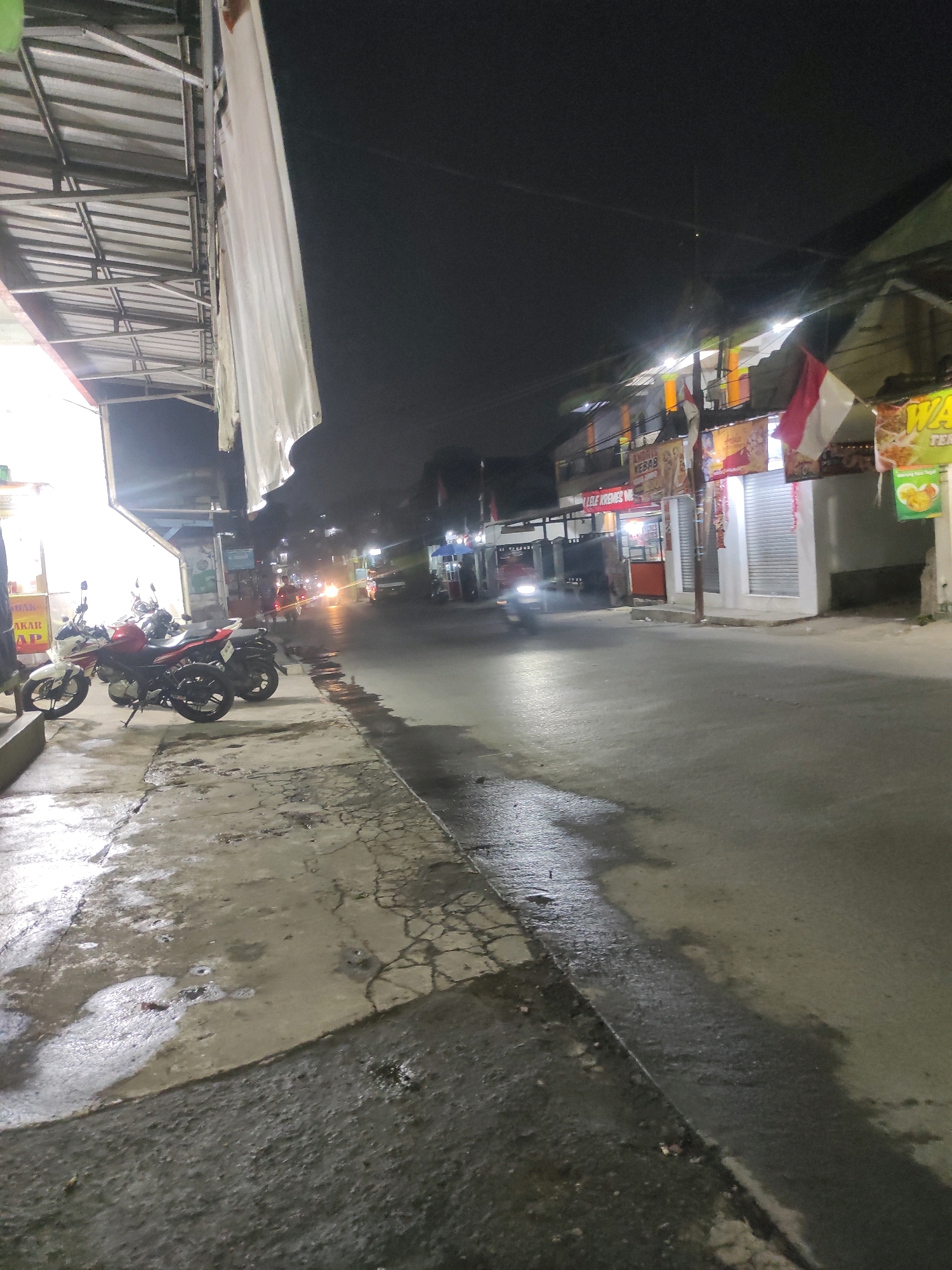 The image depicts a nighttime street scene in what appears to be an urban or semi-urban area in Padalarang, Indonesia. The foreground shows a wet, cracked sidewalk with puddles reflecting the ambient light. Several motorcycles are parked on the sidewalk. Beyond the sidewalk is a paved road with some reflections of lights, indicating it is also wet.
On the right side of the street, there are buildings with open storefronts, illuminated by artificial lights. Signs are visible above the shops, some with text and images, suggesting commercial activity, possibly food vendors. Indonesian flags, red and white, are displayed outside one of the buildings. Vehicles, including at least one motorcycle with its headlights on, are moving on the road, creating streaks of light due to the long exposure. The overall atmosphere is that of a bustling commercial street at night, possibly after rain. The background is dark, with the lights from the shops and vehicles creating pockets of illumination.