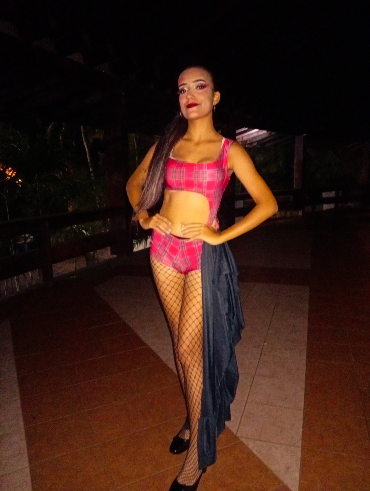 A young woman stands on a tiled surface in a dimly lit outdoor area. She is wearing a pink plaid swimsuit with a cutout at the midriff, fishnet stockings, and black flats. A dark fabric drape hangs from her waist. Her hands are placed on her hips. She has dark hair pulled to one side and wears makeup. In the background, there is a wooden railing and foliage, suggesting a balcony or patio setting. The scene appears to be in Playa El Agua, Venezuela.