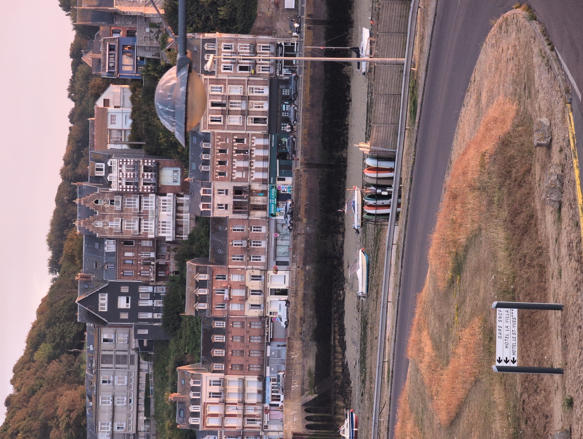 The image depicts a street scene in Le Tréport, France, featuring a waterfront promenade lined with buildings and a road in the foreground. The buildings are multi-story structures with traditional architecture, exhibiting various facades of brick and stone. The promenade is bordered by a low stone wall and a wooden fence. Several boats are visible on the exposed seabed of the harbor, indicating low tide.
In the foreground, a road curves to the right, with dry, brown grass and vegetation on either side. A signpost stands in the grass, with three directional arrows pointing left. The signs read: "GARE SNCF," "HOTEL LA VILLA," and "HOTEL LE ST-YVES***". The sky is a pale pink, suggesting either dawn or dusk. The overall scene conveys a tranquil, coastal town atmosphere.