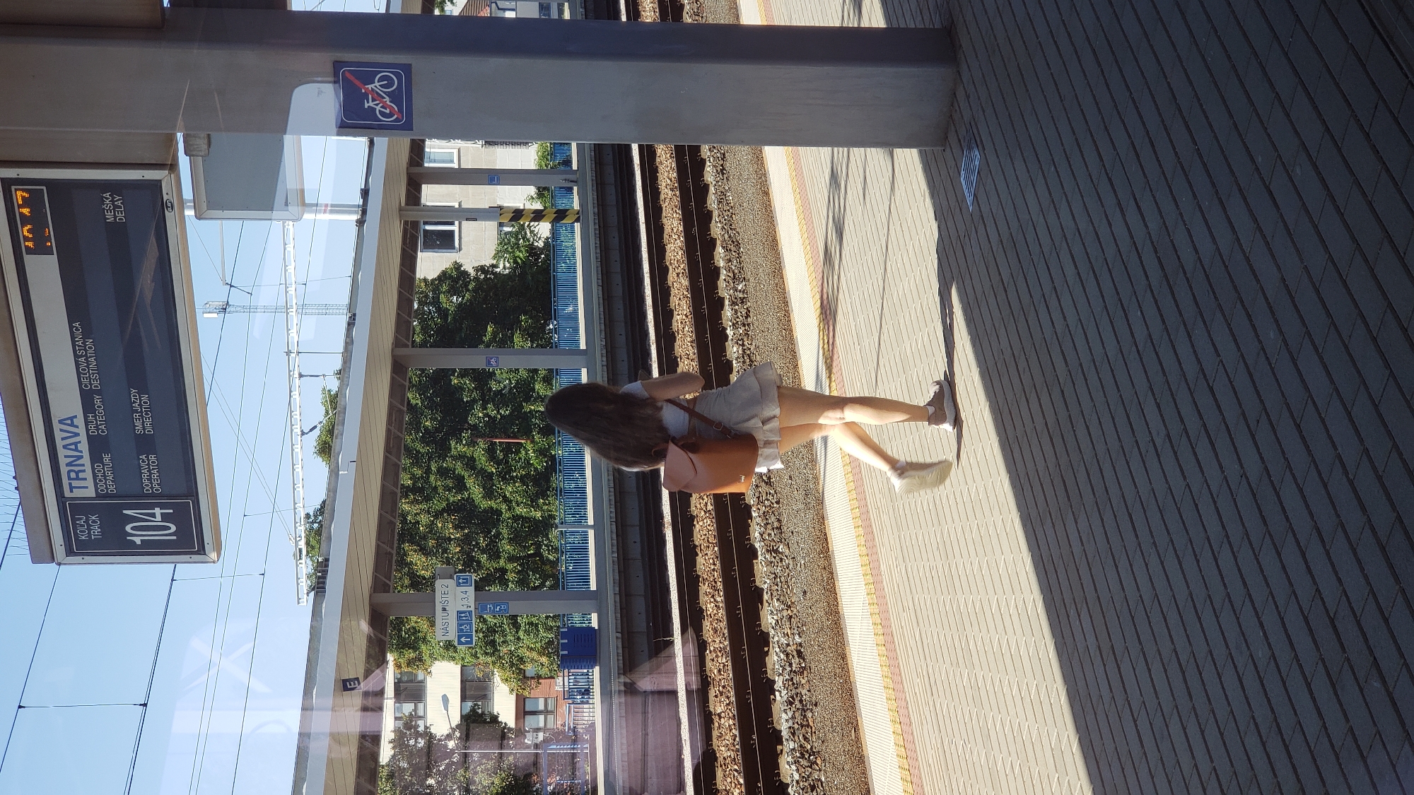 A young woman with long brown hair walks along a train platform in Trnava, Slovakia. She is wearing a light-colored skirt and top, and a brown backpack. She is walking away from the camera, towards the train tracks. The platform is paved with gray tiles and there is a shadow cast by a nearby pillar. In the background, a digital display shows train information, including "TRNAVA" as the destination. The display indicates that train number 104 is departing. The environment appears to be a sunny day, with a clear blue sky.