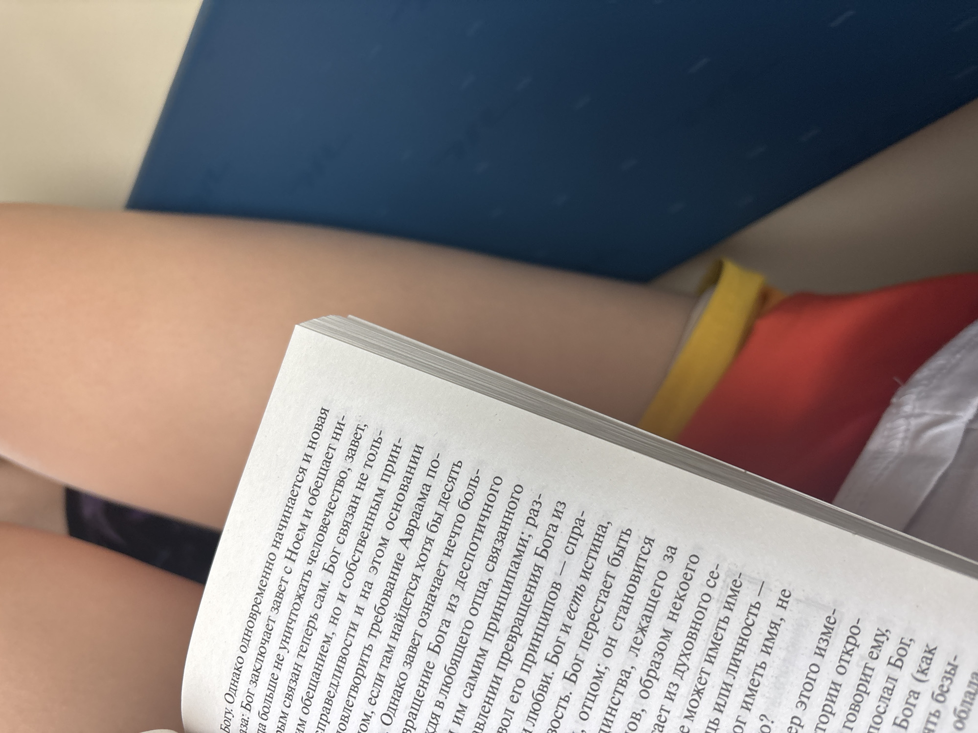 An overhead view captures the upper thighs of a person holding an open book. The book, with white pages and a visible spine on the left, displays Russian text on its right-hand page, discussing theological themes such as "God," "covenant," "Noah," and "Abraham." The person's bare skin is visible on the thighs, with a dark garment appearing on the upper left thigh and a red garment featuring a yellow band on the upper right thigh. A dark blue surface is present to the right of the book, with a lighter, beige-toned area above it, forming the background. The location in the city of Biryusinsk, Russia, is not discernible from the visual evidence within the image.