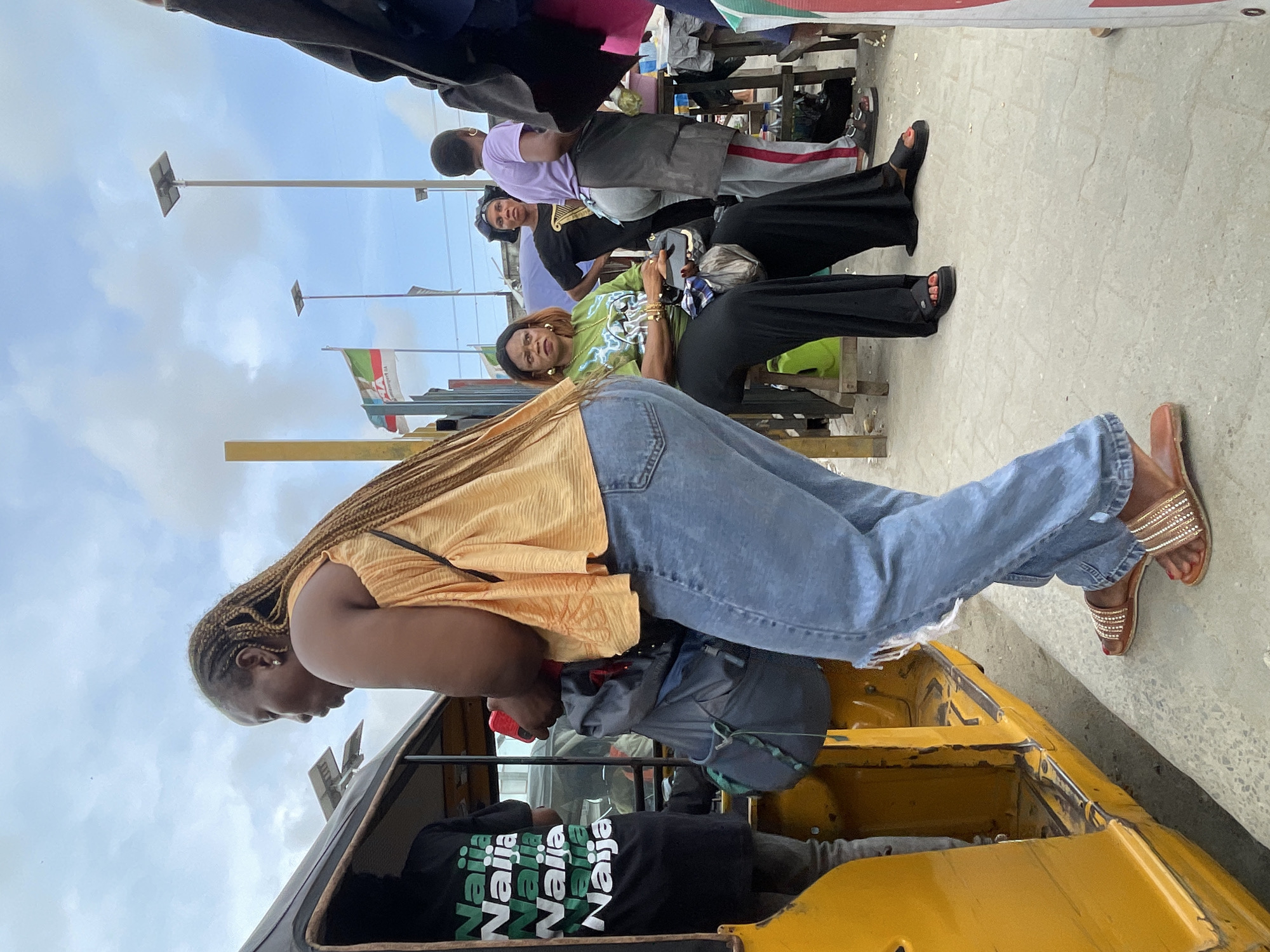 This vertical image depicts an outdoor street scene in Lagos, Nigeria, on a bright, cloudy day. In the foreground on the left, a woman with braided hair, wearing a light orange/gold top, blue jeans, and decorative flat sandals, is positioned near a yellow tricycle (keke napep). She appears to be looking into or about to enter the tricycle. Inside the tricycle, a person is visible wearing a black t-shirt with the repeating text "Naija Naija Naija Naija" printed in green and white.
To the right of the tricycle, several individuals are gathered, mostly seated on low benches or stools along what appears to be a roadside. One woman in the mid-ground, wearing a bright green t-shirt with a graphic design, black pants, and black platform sandals, looks towards the viewer. She has bangles on her left wrist. Behind her, another person in a black t-shirt and black pants is seated. Further back, another individual wears a light purple top and grey sweatpants with a red stripe.
The background shows utility poles equipped with solar panels. Several flags are visible, hanging from poles. One flag displays the green, white, and red colors, while another shows green, white, and green, typical of the Nigerian national flag, with faint text that appears to include "VP". The overall setting suggests a bustling urban environment or market area, characteristic of Lagos. The people are casually dressed, indicating a regular daytime activity.