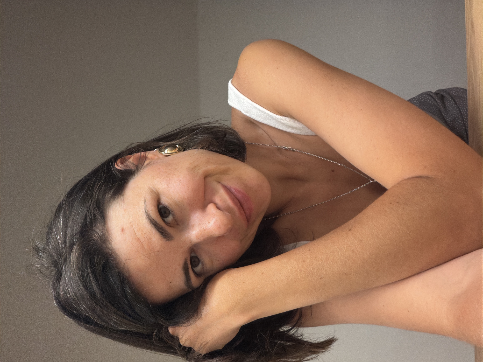 A woman with dark hair is lying down, resting her head on her hands. She is wearing a white tank top and a silver necklace. She is wearing a gold earring in her left ear. The background is a plain gray wall. The location is unknown, and there is no indication of the city of Budva, Montenegro.