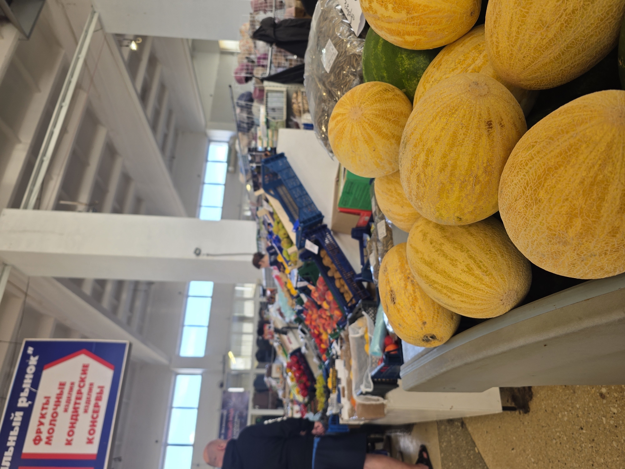 The image shows a busy indoor market in Chaikovsky, Russia, with a focus on a display of ripe melons in the foreground. The melons are a vibrant yellow and appear to be fresh and in good condition. Behind the melons, a variety of other fruits and vegetables are visible, including peaches, peppers, and what appear to be watermelons. The market stalls are organized, and customers are browsing the produce. A sign in Russian hangs in the background, indicating different sections of the market, such as "Fruits," "Dairy Products," "Confectionery," and "Canned Goods." The overall impression is one of abundance and lively commerce.
