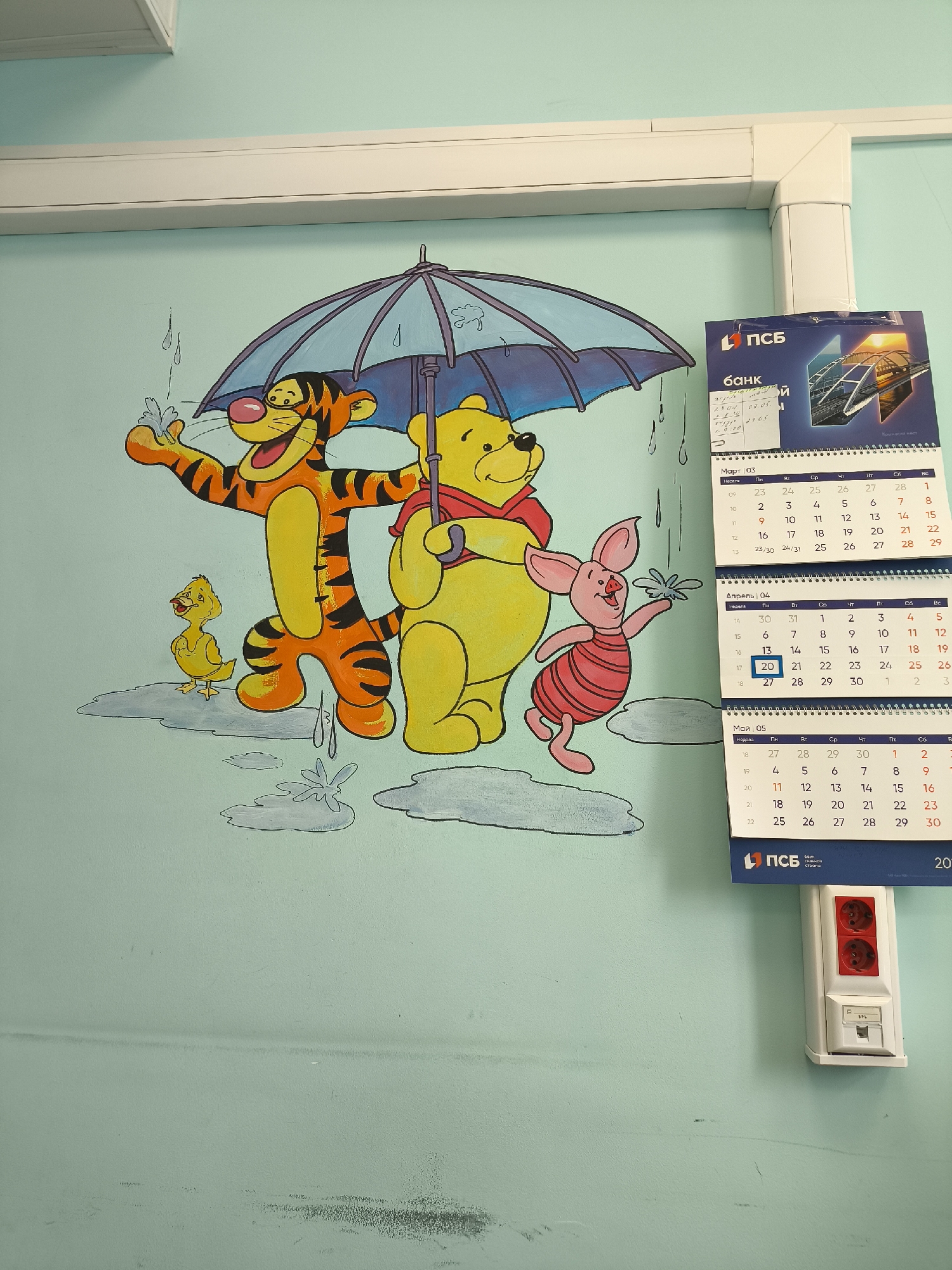 This image captures a vibrant wall mural depicting popular Winnie the Pooh characters and other objects in an indoor setting, likely in Khimki, Russia. The mural features Tigger, Winnie the Pooh, Piglet, and a small yellow duckling, all enjoying what appears to be a rainy day under a shared blue umbrella. Tigger is on the left, depicted with a wide, joyful grin, splashing water with one hand. Winnie the Pooh stands centrally, holding the umbrella handle with a calm expression, wearing his characteristic red top. Piglet, to Pooh's right, is shown mid-step with an excited look, holding a splash of water or a dandelion. The ground is represented by grey puddles and water splashes, reinforcing the rainy theme.
Above the mural, a white cable trunking system is visible, running along the wall and corner, possibly housing electrical or data cables. To the right of the mural, a stack of three tear-off calendars hangs on the wall. The top calendar displays the logo "ПСБ БАНК" (PSB BANK) and an image of a bridge at sunset. Below it, the months of March, April, and May are visible, with dates and days of the week in Russian. April 20 is highlighted on the April calendar. A sticky note on the top calendar has handwritten dates like "23.04", "07.05", and "21.05". Below the calendars, a white electrical socket unit with two red circular outlets and a white data port (labeled "SPL") is mounted on the wall. The overall scene suggests a cheerful and welcoming environment, possibly in a public space or a children's area, given the mural's subject matter.