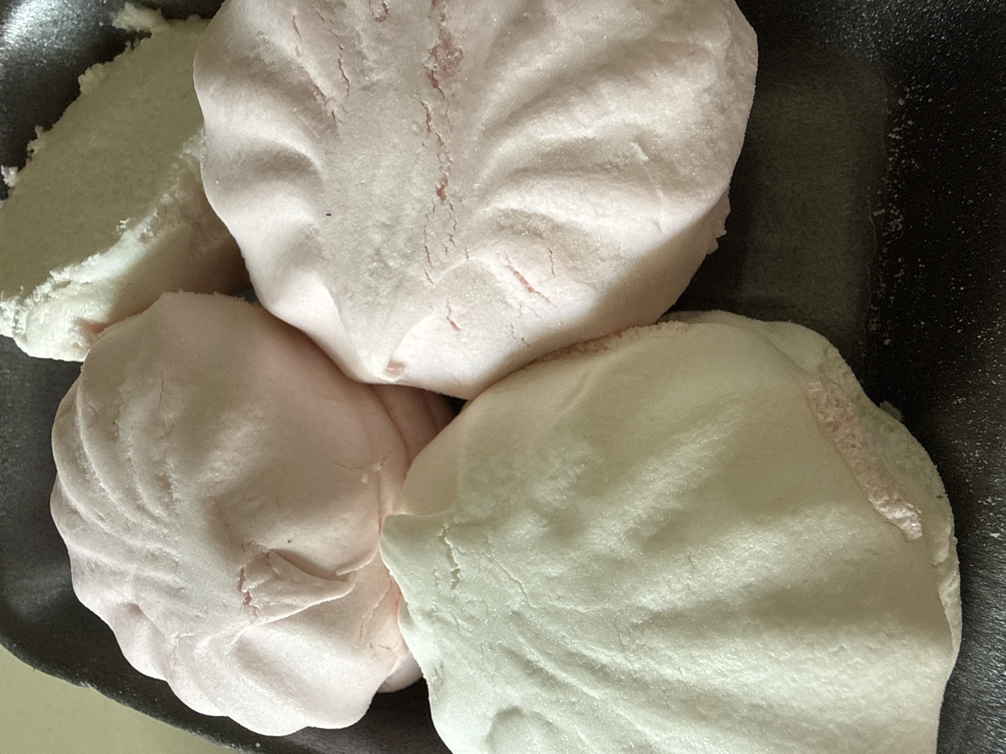 A close-up, overhead shot shows four large, light pink and off-white marshmallows arranged in a dark gray, rectangular plastic tray. The marshmallows are soft and appear to have a slightly textured, powdery surface with visible creases and subtle cracks. Some of the pink marshmallows exhibit a lighter pink interior peeking through. The tray itself is standard for packaging baked goods and is filled with a dark, slightly gritty residue, particularly in the lower right portion of the frame. The overall lighting is soft and diffused, highlighting the gentle contours of the marshmallows. The image is likely a product photograph, emphasizing the texture and form of the confectionery.