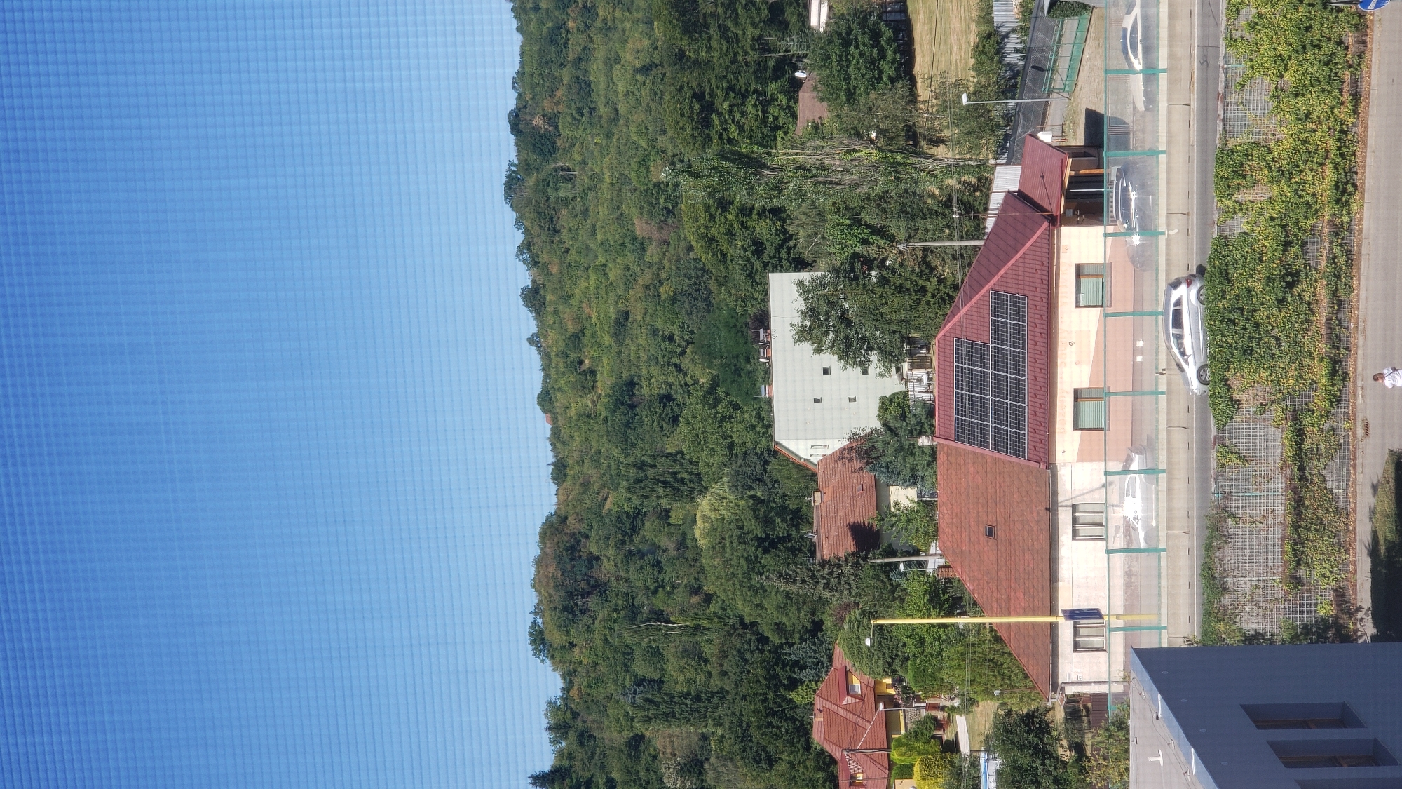 The image depicts a view of a residential area with a wooded hillside in the background, likely on the outskirts of Prešov, Slovakia. In the foreground, a multi-story building with a grey facade is visible on the left. A road runs horizontally across the middle of the frame, with several cars driving on it. On the right side of the road, a house with a red tile roof and solar panels is prominent. Beyond the houses, a dense green forest covers a rolling hill, suggesting a semi-urban or suburban setting with proximity to nature. The sky above is a clear, bright blue.
