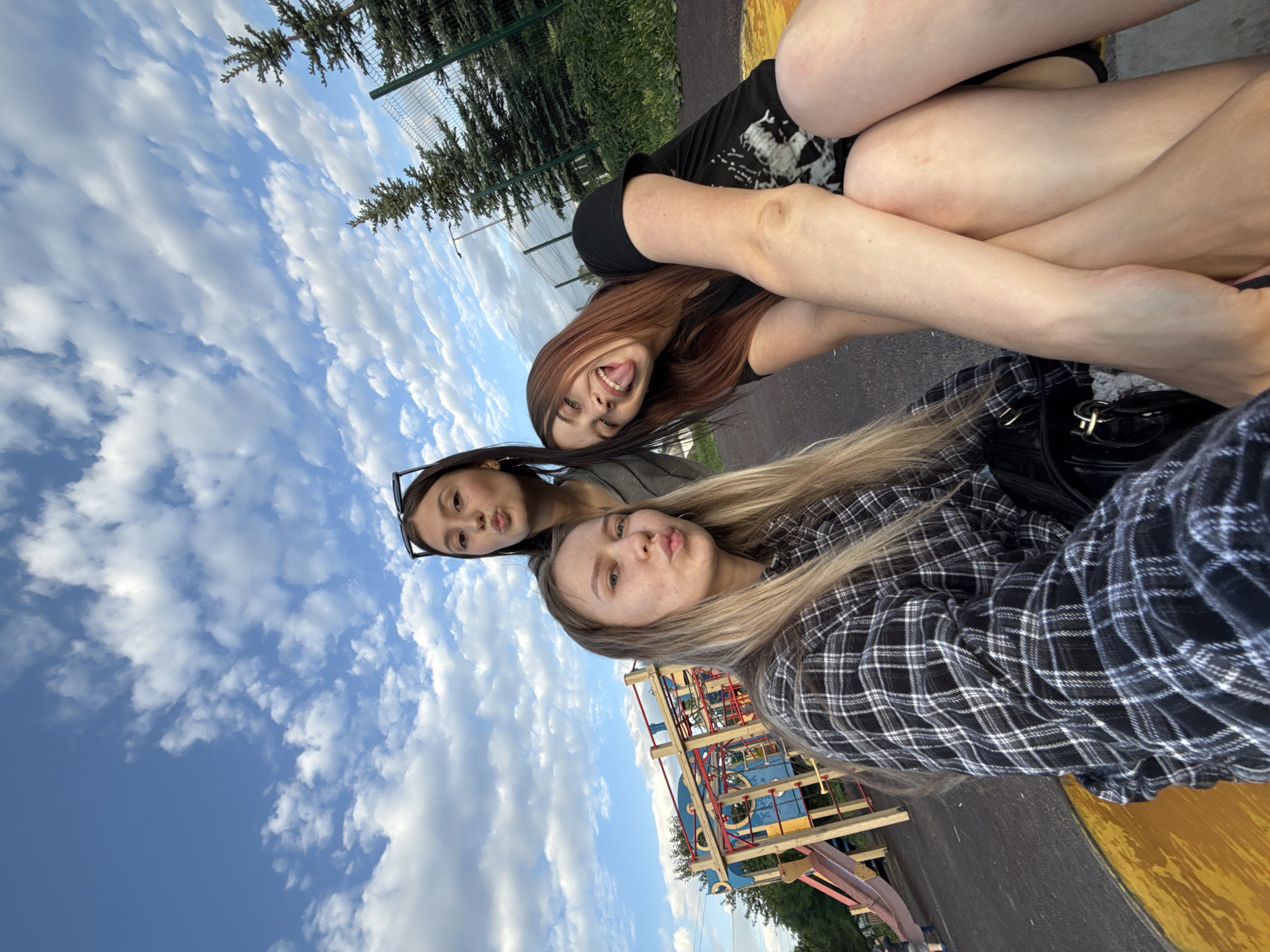 Three women are posing for a photograph outdoors. One woman is sticking her tongue out. Another is wearing glasses and pursing her lips, while the third woman is also pursing her lips. A playground is visible in the background. The location is Фоки, Russia.