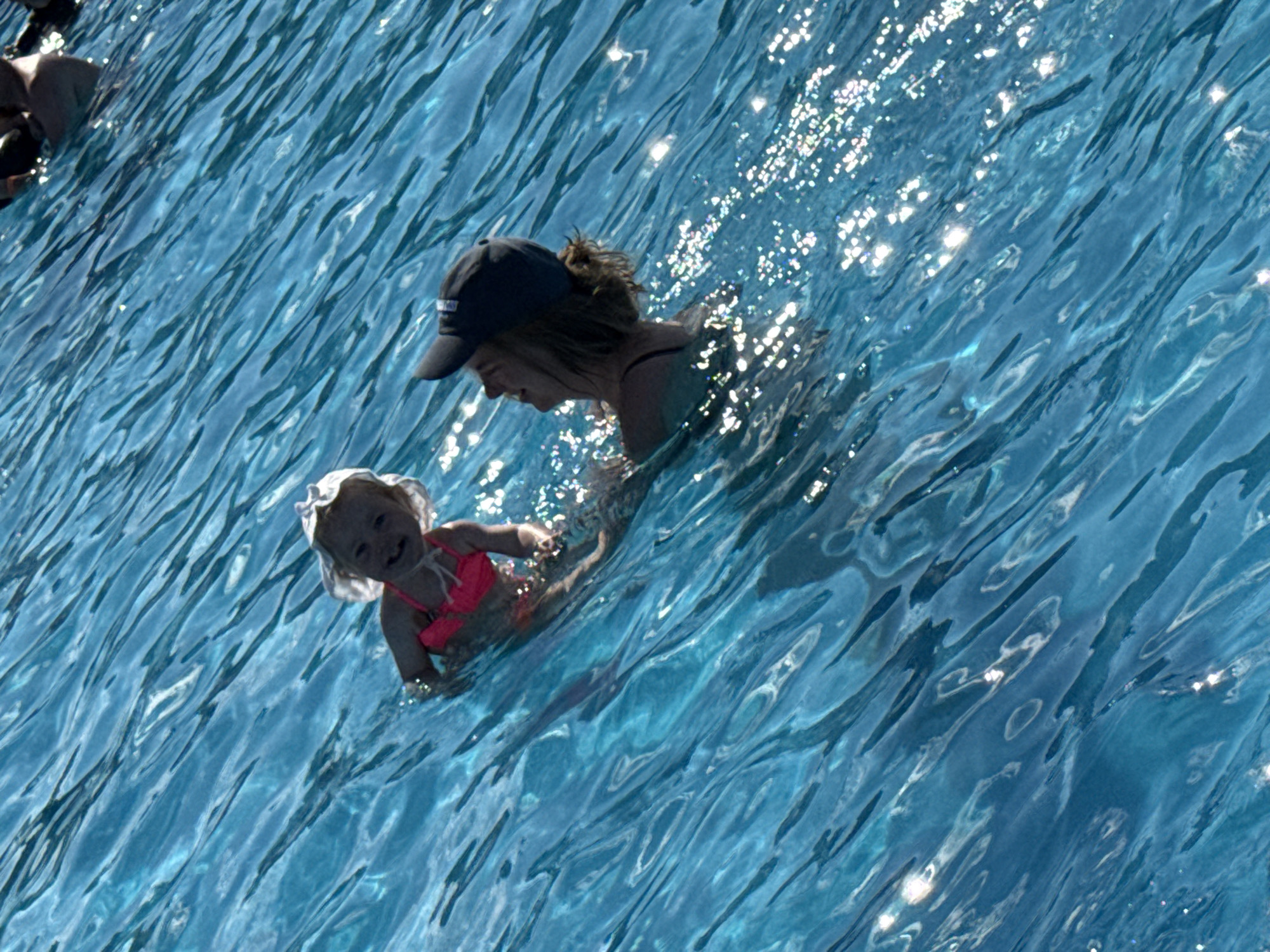 Two individuals are in a body of bright blue water. On the left, a young child wearing a pink swimsuit and a white sun hat floats on their back, looking towards the viewer with a smile. To the right of the child, a person wearing a dark baseball cap and a dark swimsuit is seen from the side, looking up and smiling. The water is slightly rippled with sunlight reflecting off the surface. The scene appears to be outdoors, possibly in a swimming pool or a calm body of water. There is no visible indication of the city of Угор, Russia.