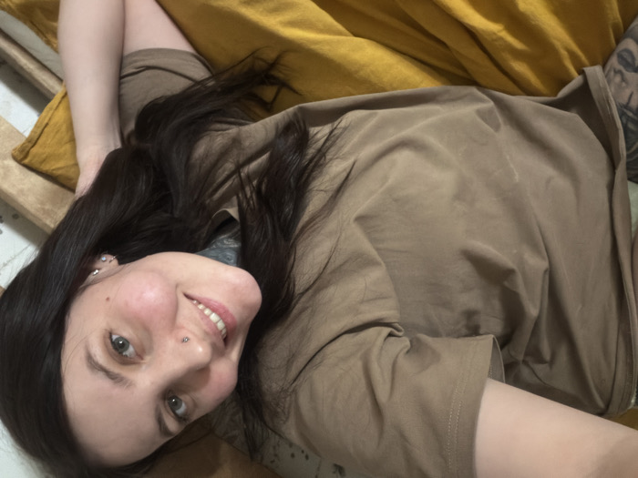 A female subject is depicted in a diagonal selfie, lying supine on a bed in an indoor residential setting within the city of Chaikovsky, Russia. The individual has dark hair, green eyes, and a fair complexion. A silver stud piercing is visible on her left nostril, and multiple small piercings are present on her visible right ear. She is smiling towards the camera, revealing her teeth. The subject is wearing a plain, loose-fitting, light brown short-sleeved t-shirt. Tattoos are visible on her left arm, extending from beneath the sleeve, and partially on her neck/shoulder area, obscured by hair and clothing. She rests on a mustard yellow colored sheet or duvet cover. A light-colored wooden bed frame or headboard is visible on the right side of the image, and a subtle, lighter-toned patterned surface is present in the upper right background.