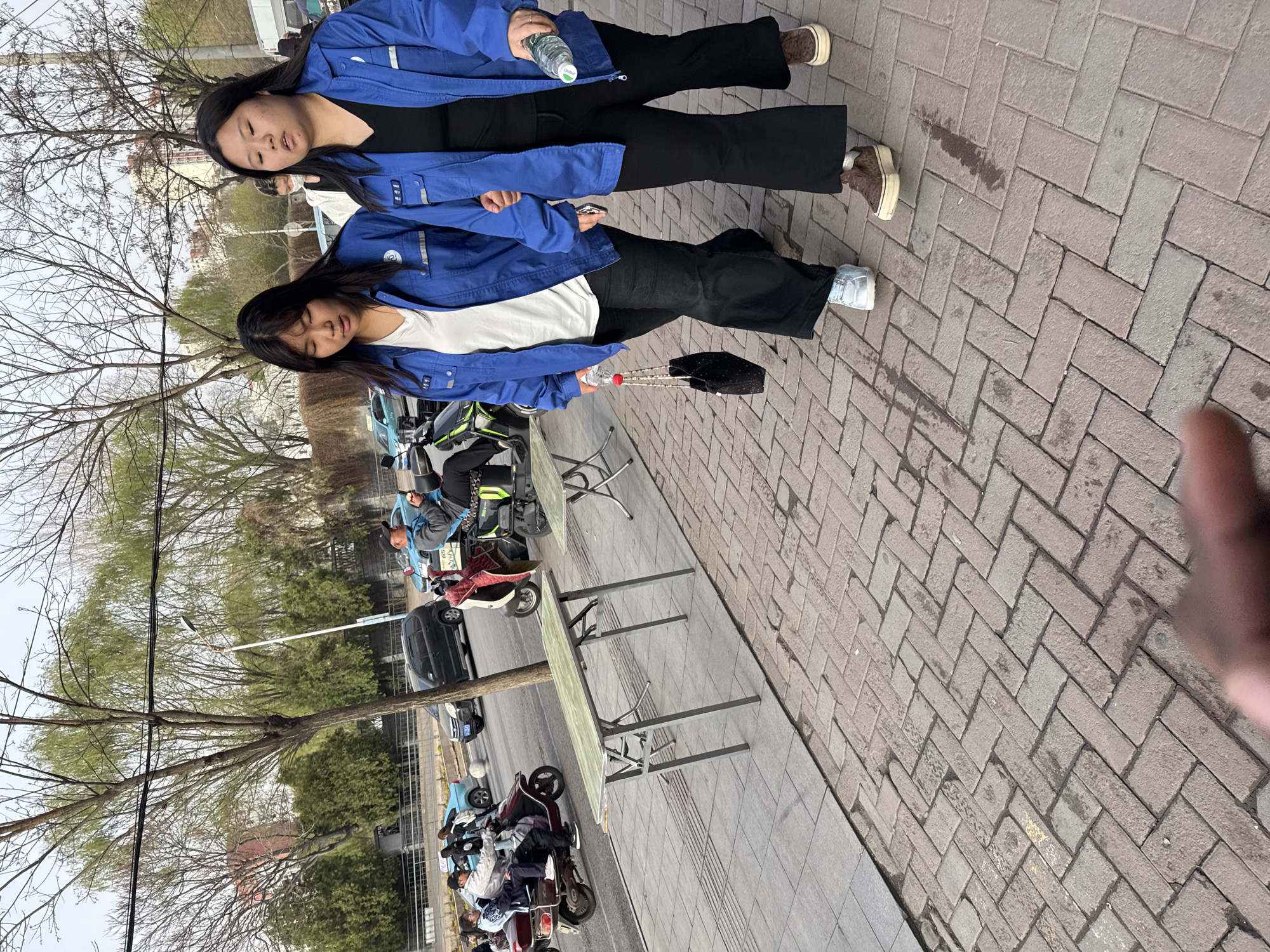 Two women in blue jackets stand on a brick sidewalk in Zhongtun, China. One woman wears black pants, a black shirt, and brown shoes, holding a water bottle. The other woman wears black pants, a white shirt, and white sneakers, carrying a black purse and water bottle. Behind them, scooters are parked along the street next to folding tables. A finger is visible in the lower right corner. A tree stands along the sidewalk, and a street with cars is visible in the background.