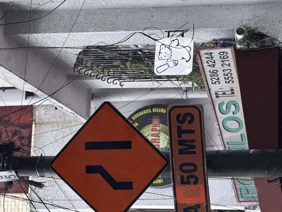 An urban street scene in Mexico City, Mexico, features a densely wired utility pole in the foreground against a textured, light-colored building facade. The utility pole is dark and heavily entangled with numerous black electrical and communication wires near its top.
Attached to the pole, from top to bottom, are three signs:
1. A diamond-shaped orange traffic sign with a thick black border, displaying two black vertical lines, one straight and one angled from the left, indicating a left lane merge or narrowing.
2. Below it, a rectangular brown sign with a black border, showing "50 MTS" in black letters, signifying "50 Meters." A faded orange box partially obscures text preceding the numbers.
3. Further down, partially visible behind the brown sign, is a circular sign with a yellow center and black border, displaying text in Spanish that includes "BORDADOS," "DISEÑO," and "GRAPHICS."
The building facade to the right is textured, likely stucco or concrete, and appears light gray or off-white. An arched window opening is visible, secured by a dark, ornamental metal grille through which green foliage grows.
Affixed to the building are two additional signs:
1. A square white sign with a black outline, featuring a stylized, cartoonish white bear holding a chainsaw, depicted with motion lines.
2. Below the bear sign, a rectangular sign with a white background and green lettering displays "TEL: 5286 4244" and "5553 2169," indicative of telephone numbers. The beginning of a Spanish word, possibly "CULOS" or part of a longer word, is visible in green before the phone numbers.
Numerous black wires crisscross the entire scene, extending from the utility pole towards the building and beyond. A white PVC pipe protrudes from the building's facade below the telephone number sign, with green foliage growing around its base.