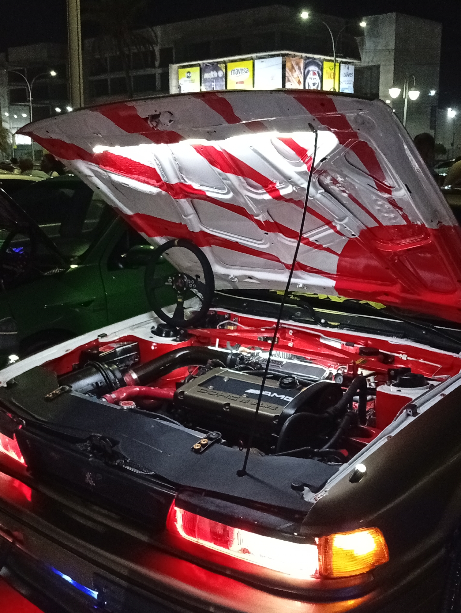 The image shows the engine bay of a modified car at night. The car's hood is open and painted with a red and white rising sun pattern. The engine is visible, along with a steering wheel. The surrounding environment appears to be a street or parking lot in Porlamar, Venezuela, as suggested by the "Mavesa" signs in the background, a brand commonly found in Venezuela. Streetlights illuminate the scene. Another car is visible on the left. There are people visible in the background, but they are out of focus.