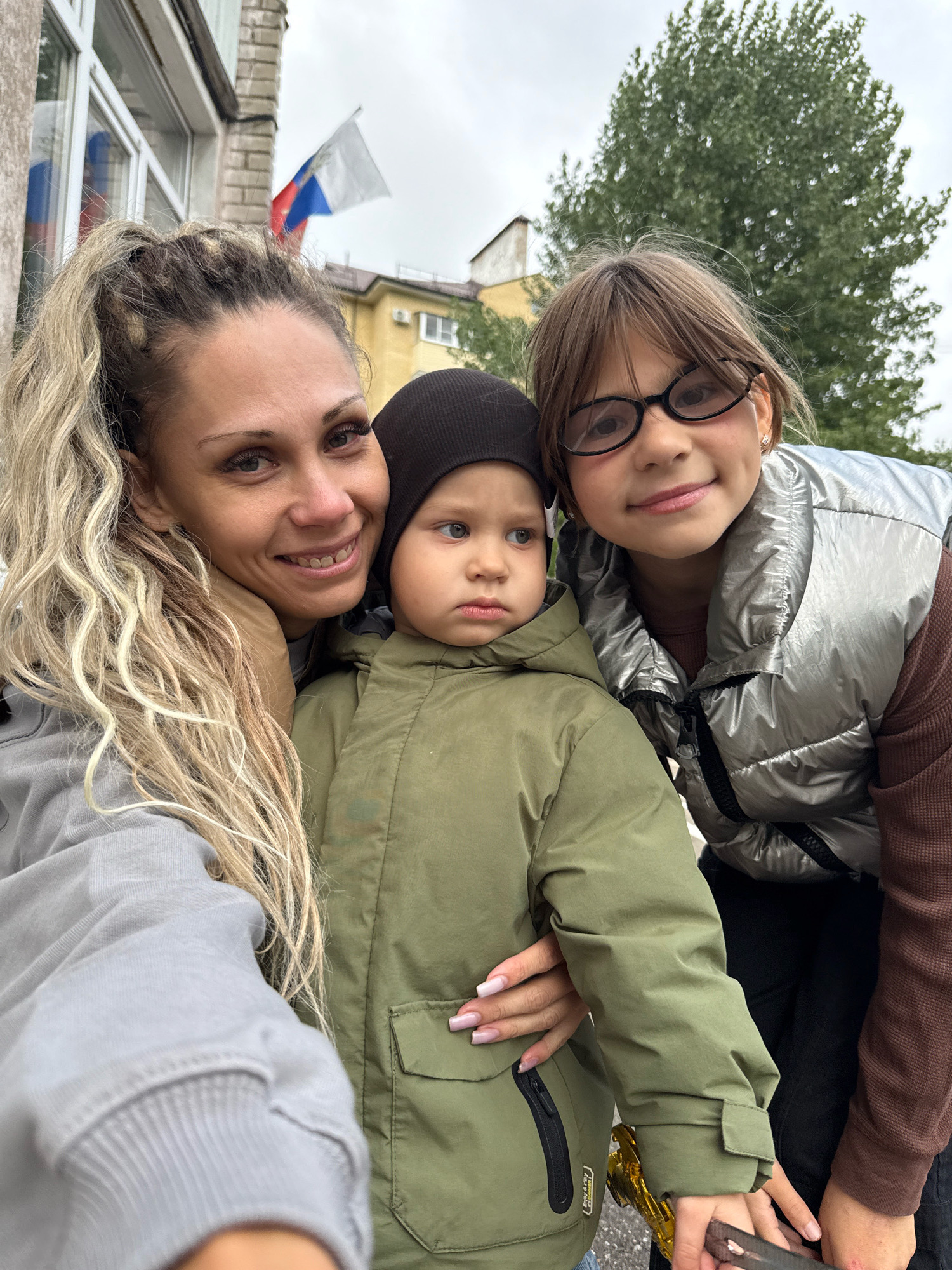 In Rusanovo, Russia, a woman with blonde, braided hair is taking a selfie with two children. The woman is on the left, smiling and leaning in towards the camera, with her arm around the younger child. The younger child, a boy wearing a black beanie and a green jacket, is in the center, looking slightly to the right. To the right of the younger child is an older girl wearing glasses and a silver puffer vest over a brown shirt. She is also looking towards the camera. In the background, there is a building with a Russian flag flying. A large tree is visible behind the children and the building. The scene appears to be outdoors on an overcast day.
