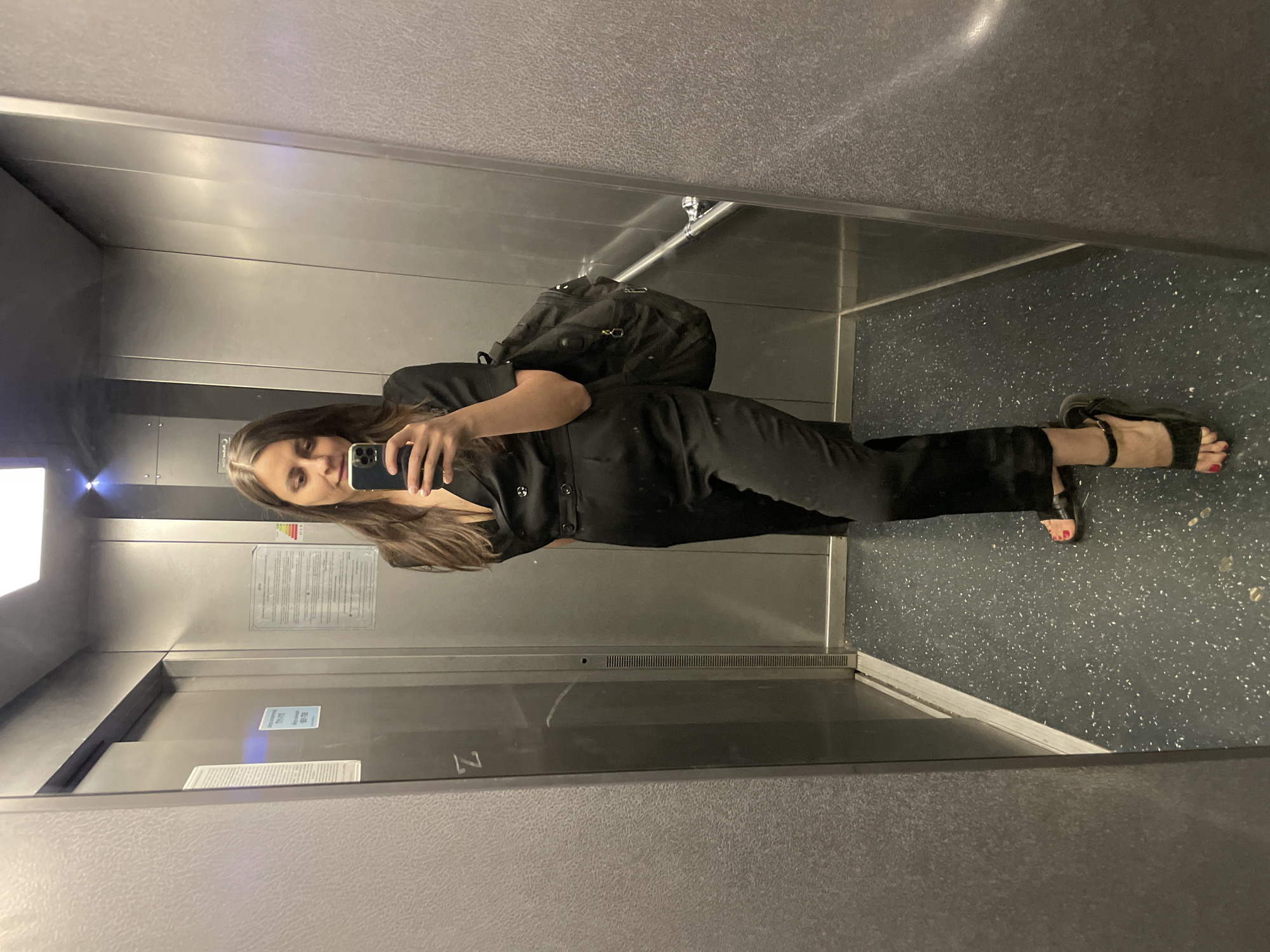 A woman is taking a selfie in an elevator. She is wearing a black jumpsuit and sandals, and carrying a black backpack. The elevator is silver and gray, and appears to be in a building with multiple floors. The location is Unknown, Unknown.