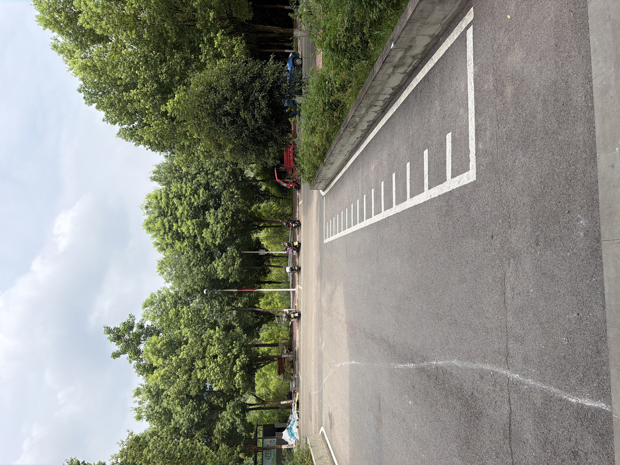 The image shows an outdoor paved area, likely a parking lot or road, with a concrete barrier and lush green vegetation along the right side. White painted lines on the pavement suggest designated parking spaces or a driving lane. In the background, a line of trees forms a dense green canopy under a partly cloudy sky. Several parked motorcycles are visible near a sidewalk, along with some piles of materials and debris. A red three-wheeled vehicle and a blue car are also present. The scene appears to be in a park-like or semi-urban setting within Jiangjin, China, given the presence of these vehicles and the surrounding vegetation. The overall impression is a quiet, functional outdoor space.